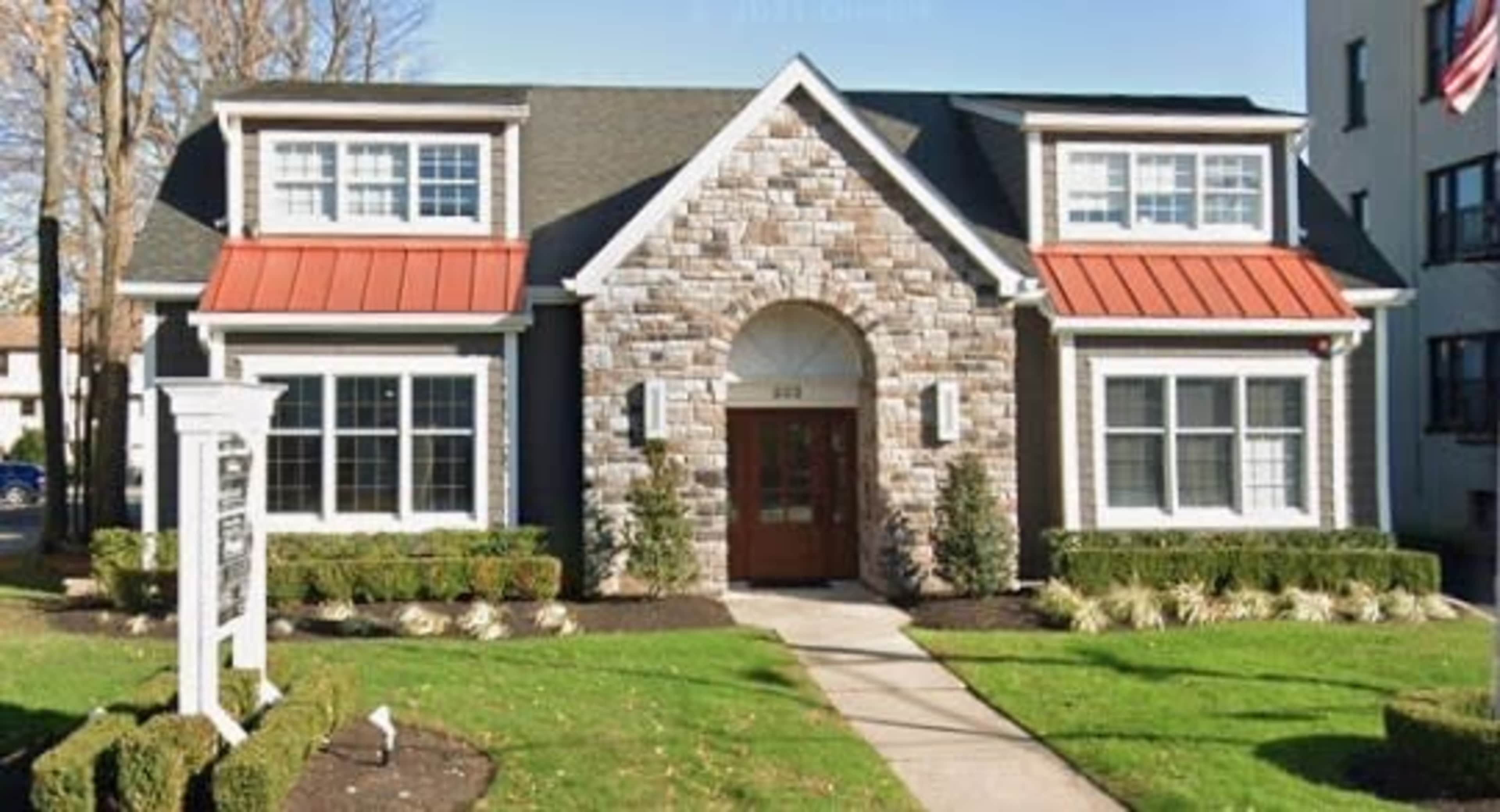The image shows a two-story building with a stone facade, red metal roof accents, and large windows set in a landscaped yard.