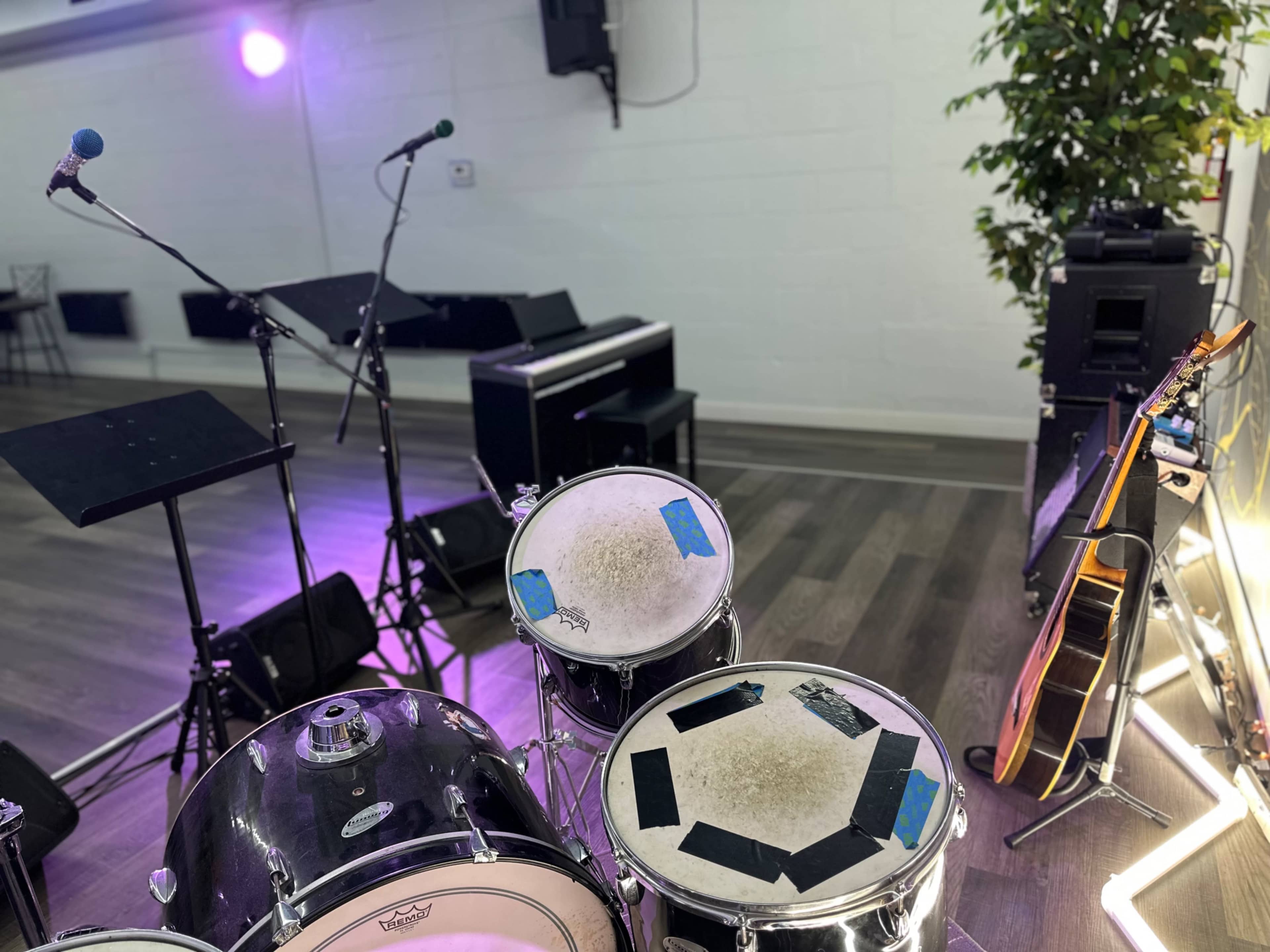 The image shows a stage setup with a drum kit, microphones, a keyboard, amplifiers, and a guitar, surrounded by potted plants and ambient lighting.