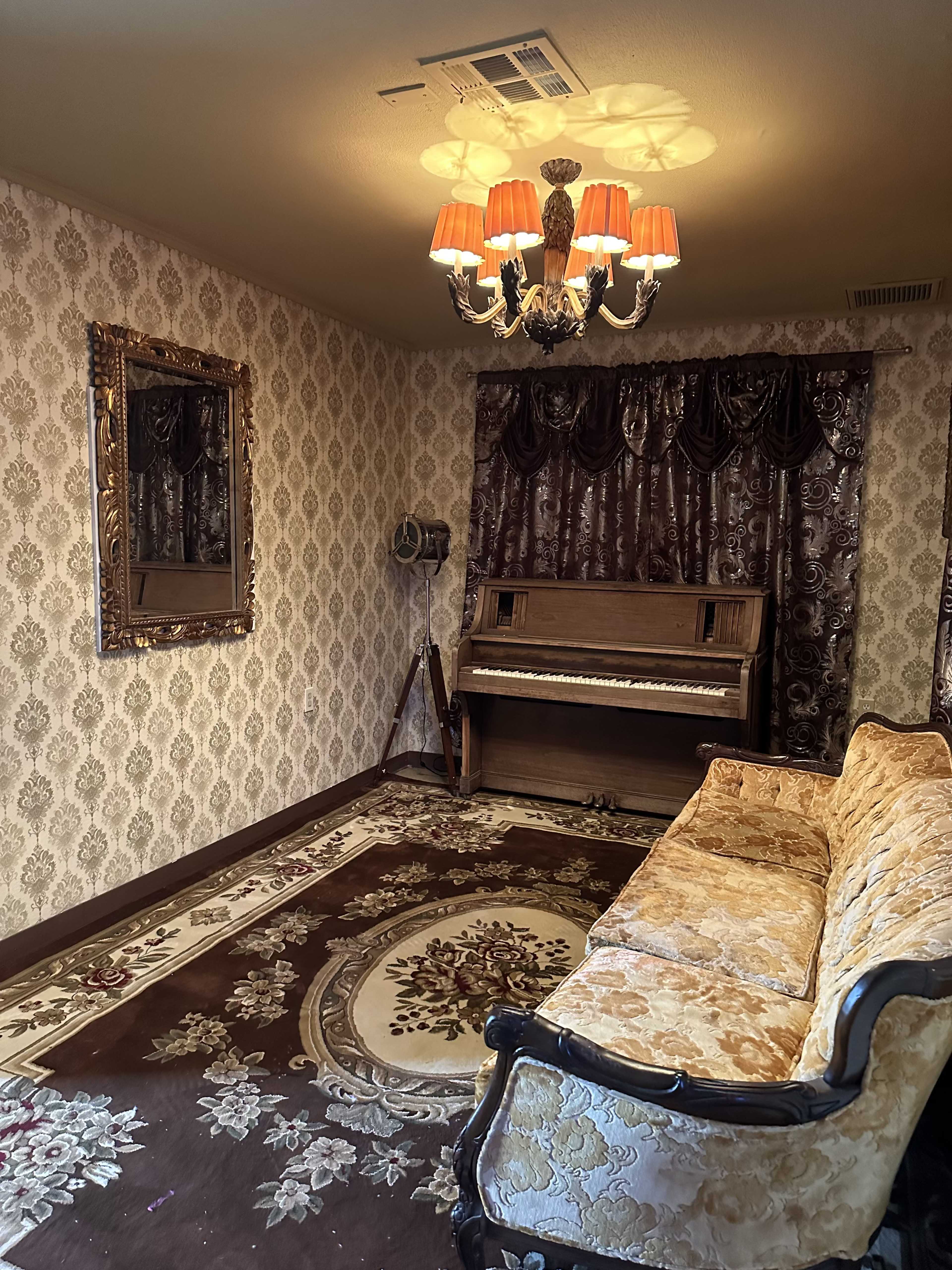 The room features a vintage piano, a patterned rug, an ornate mirror, and a chandelier, with upholstered seating against a floral wallpaper background.