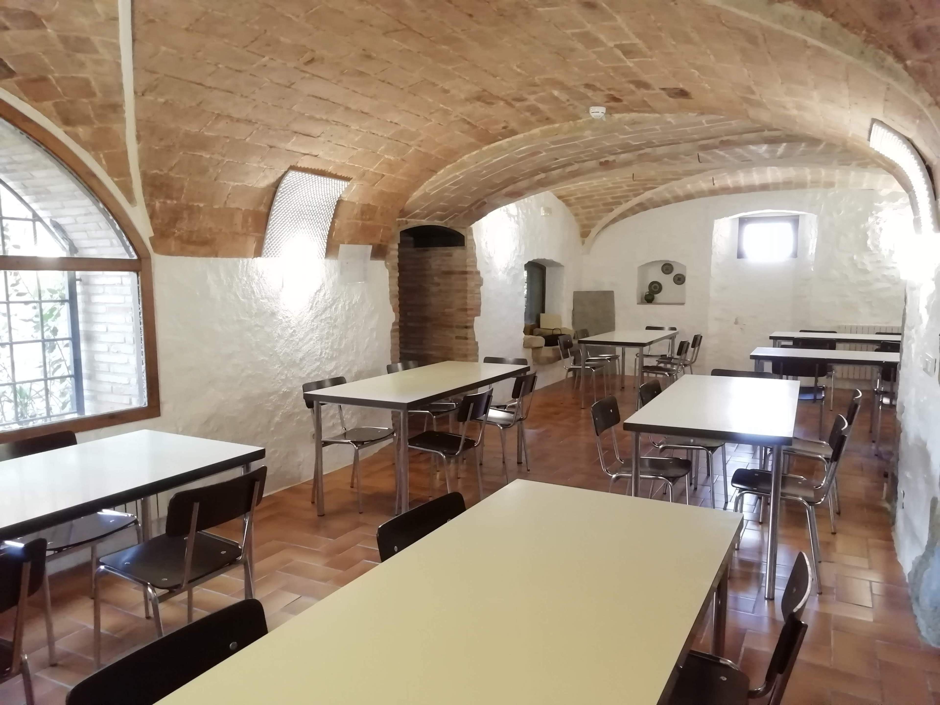 The image shows a spacious, brick-vaulted room with several rectangular tables and chairs arranged throughout.