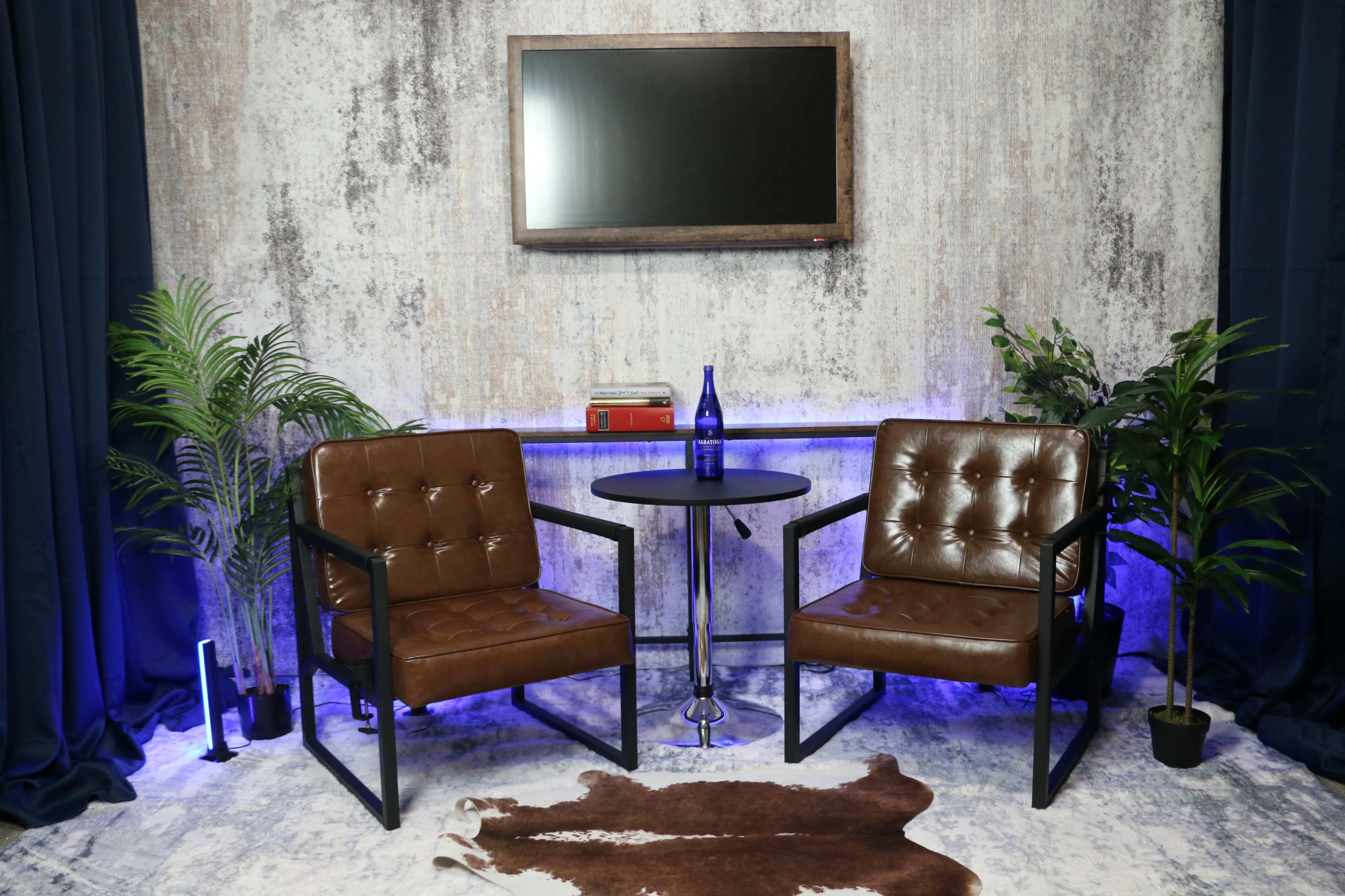 The image shows a styled seating area with two brown armchairs, a small round table, a blue bottle, a stack of books, and decorative plants against a textured wall and dark curtains.