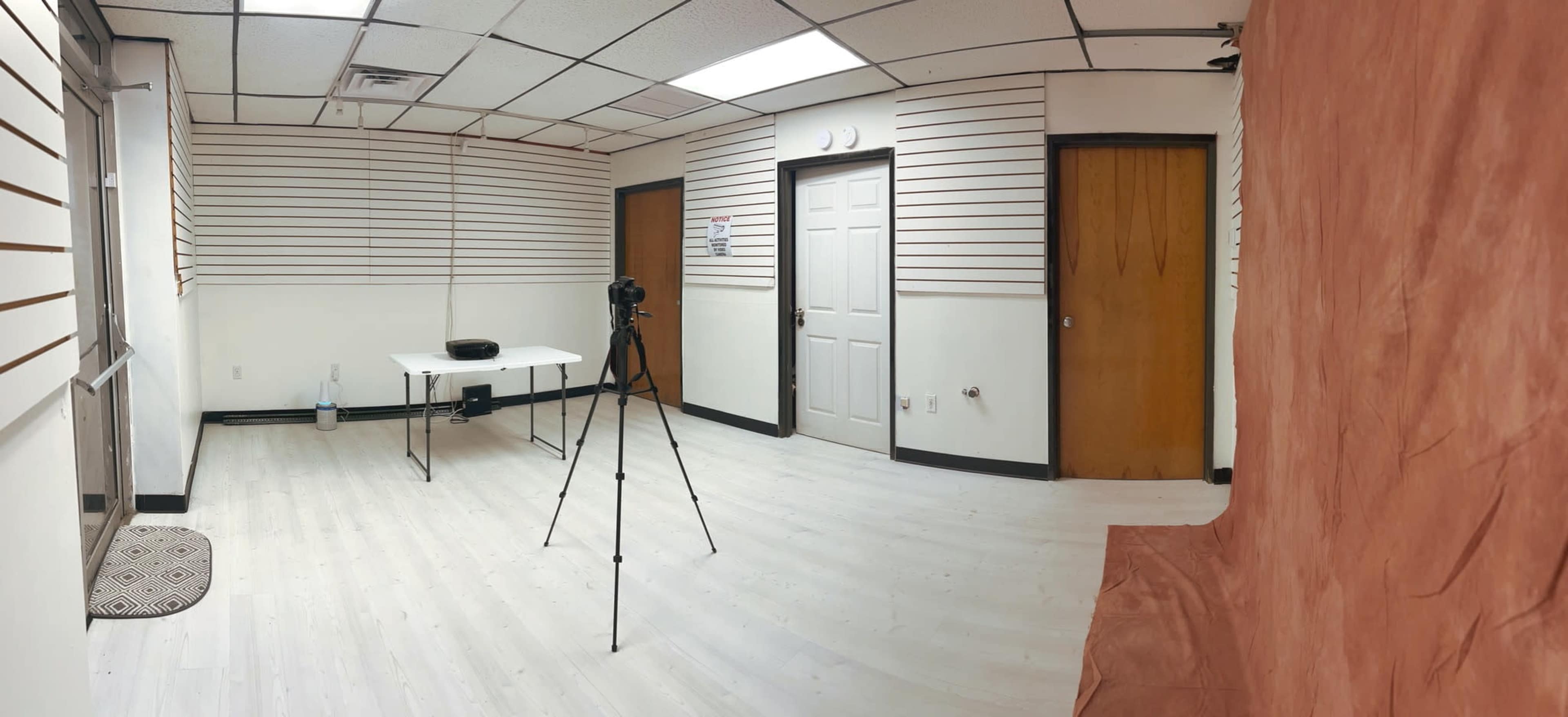 A panoramic view of a sparsely furnished room with a white floor, a table and camera setup, and plain walls featuring horizontal lines.
