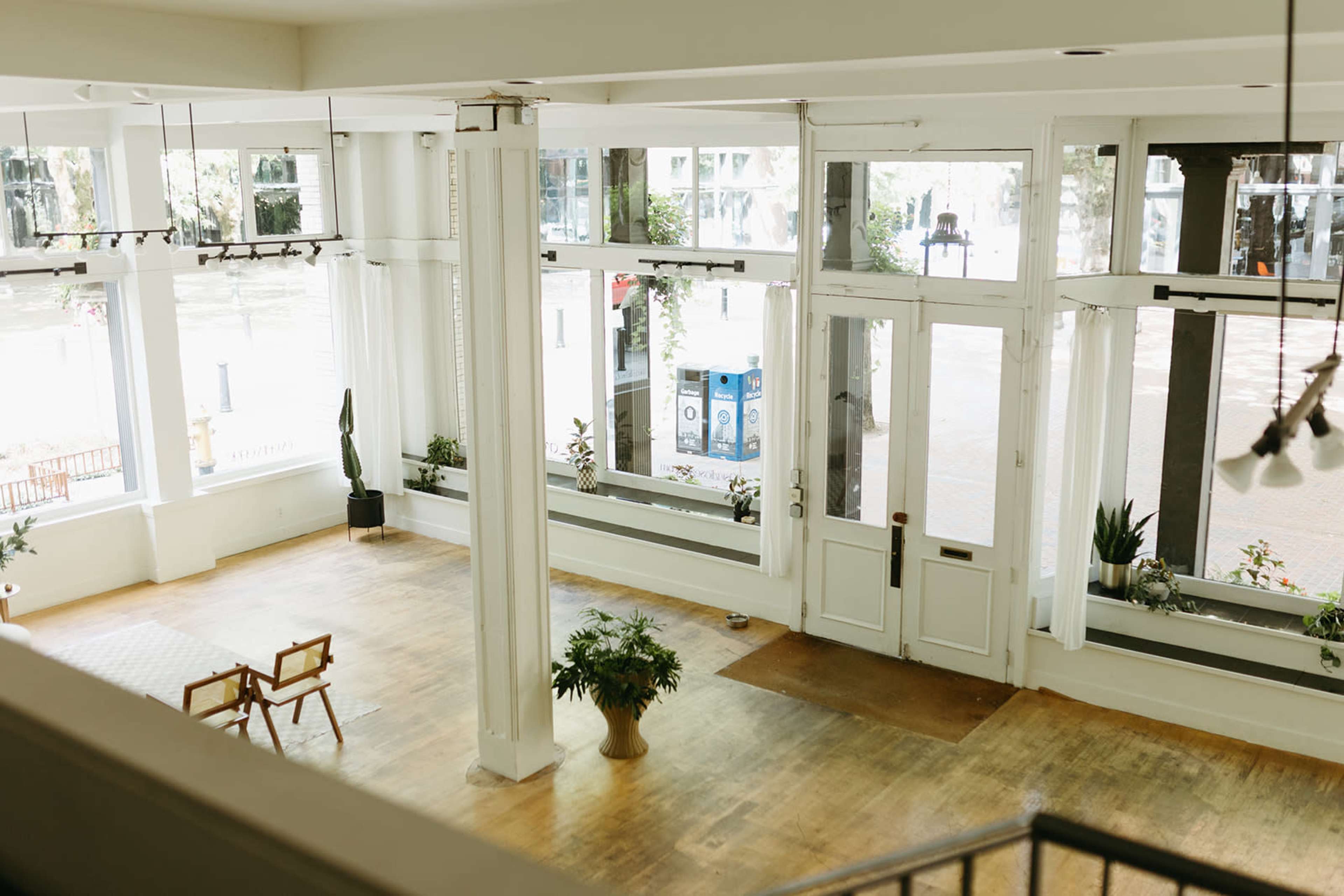 The image depicts a spacious, well-lit room with large windows, wooden flooring, and minimal furniture, including a couple of chairs and potted plants.