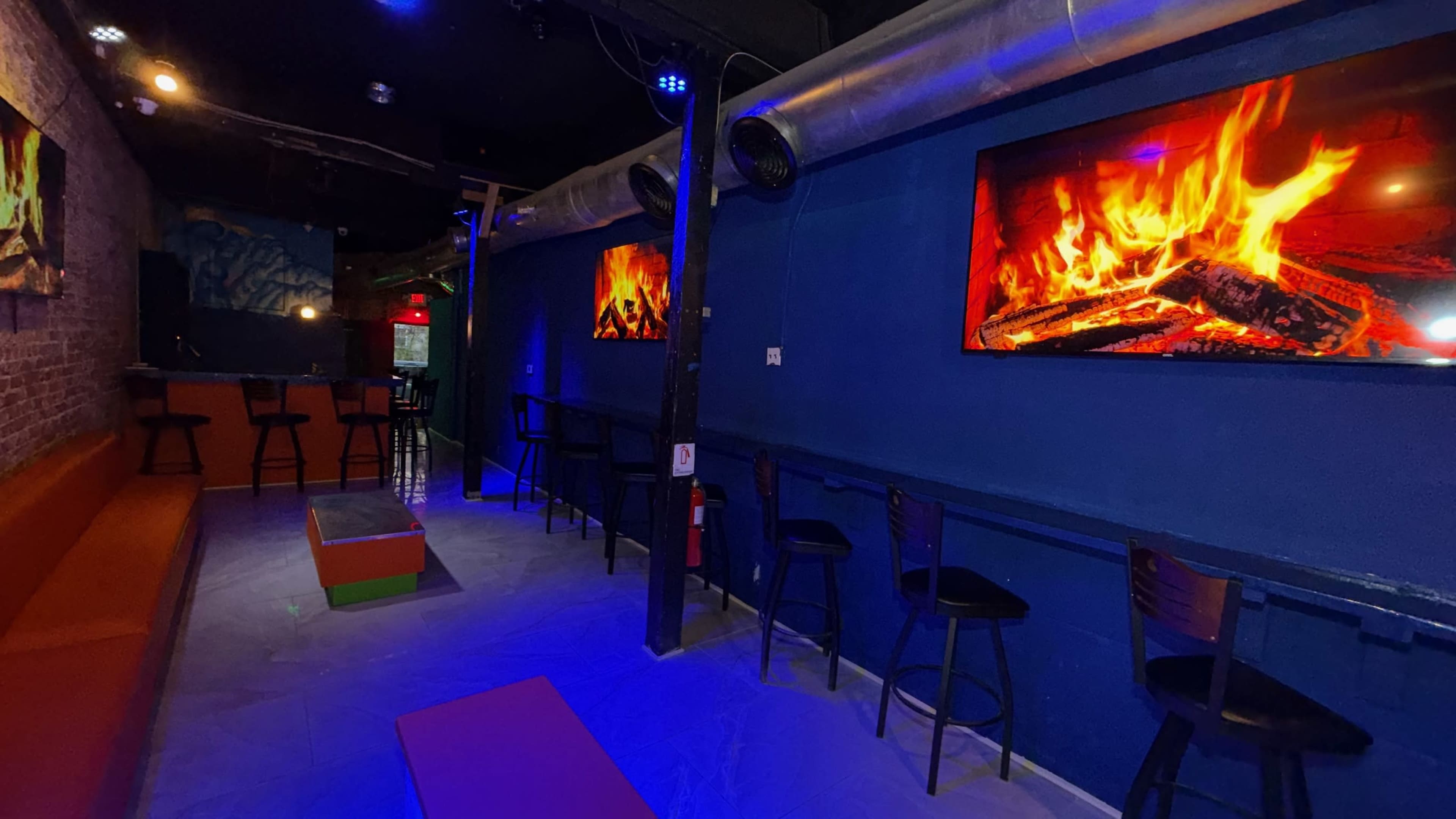 The image shows a dimly lit bar setup with colorful LED lights, featuring two large screens displaying animated fires, high chairs along the walls, and a bar counter in the background.