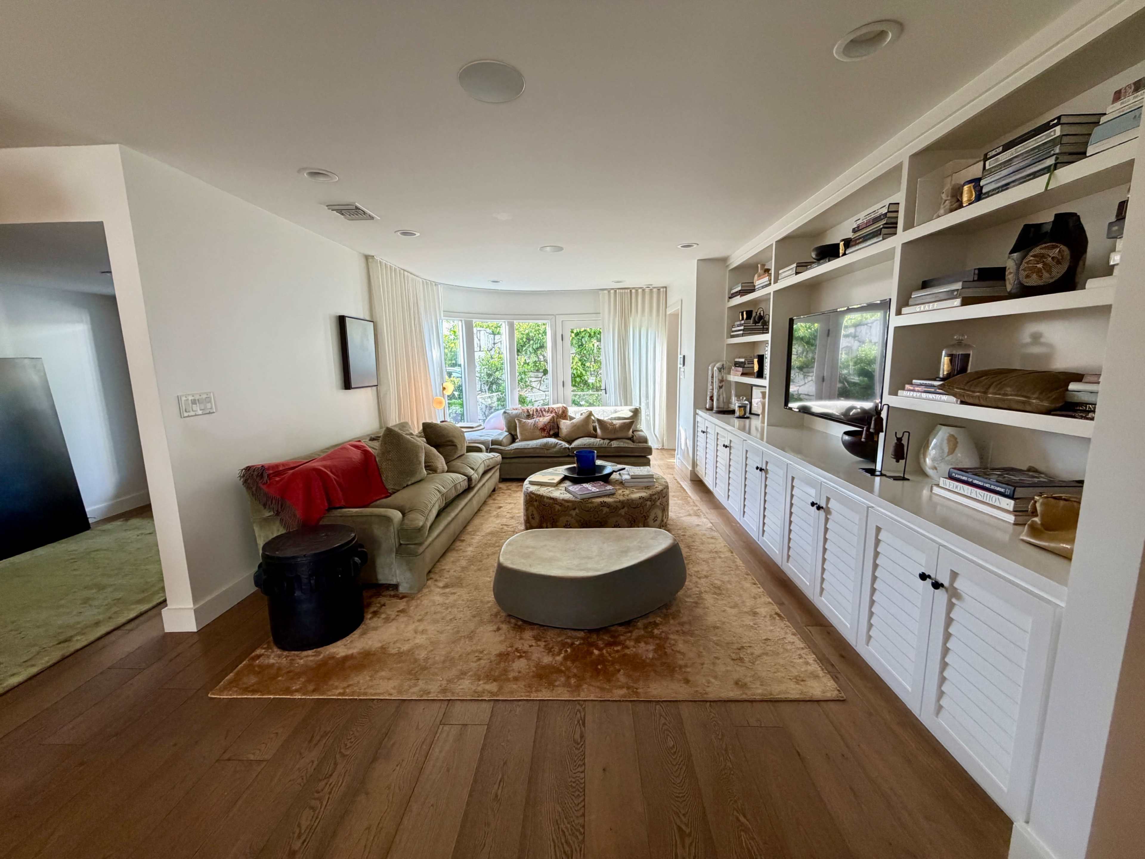 The image shows a modern living room with a large sofa, an ottoman, built-in shelves filled with books and decorative items, and large windows allowing natural light.