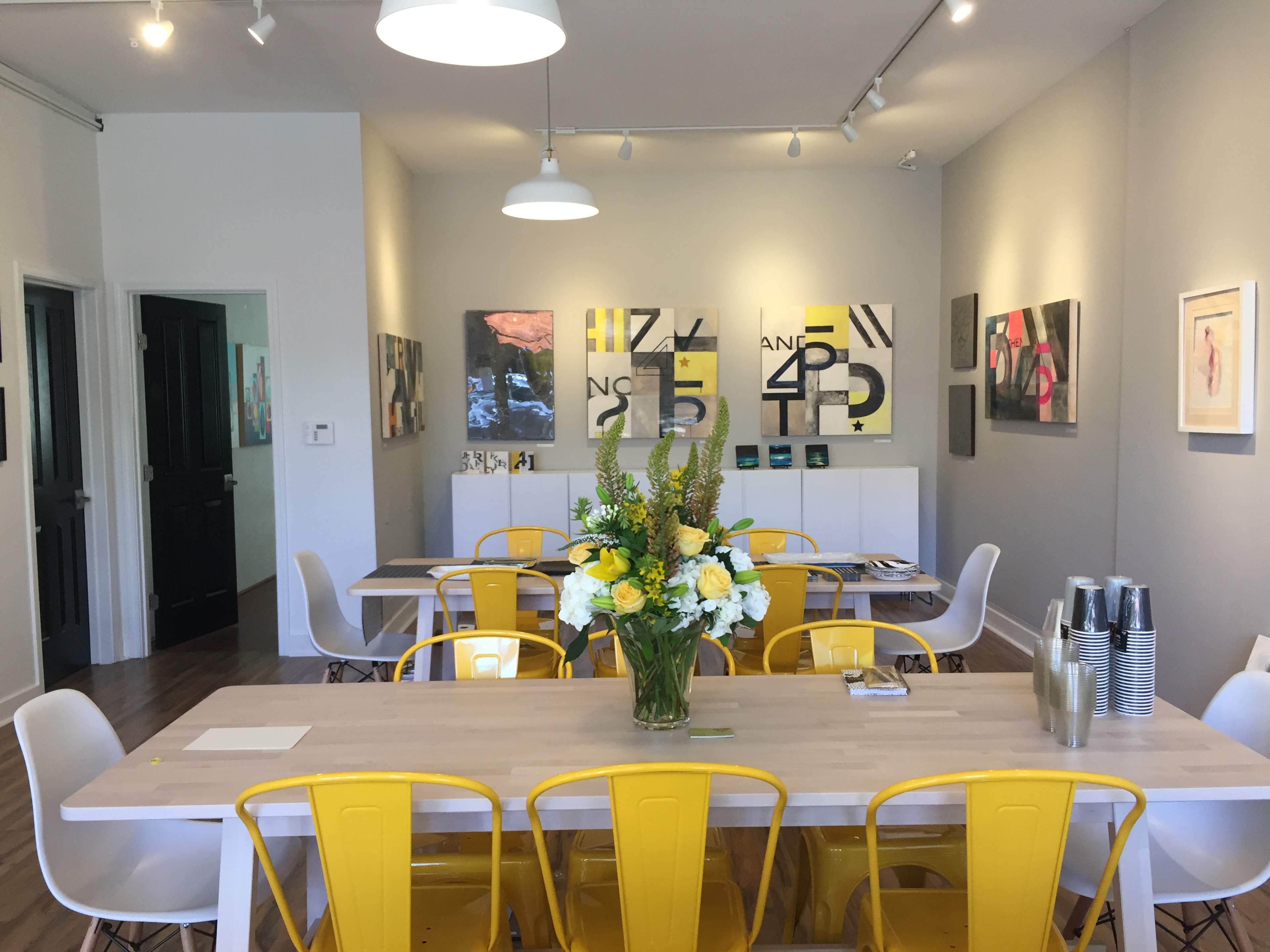 A dining area features a long wooden table surrounded by yellow and white chairs, with artwork on the walls and a floral centerpiece.