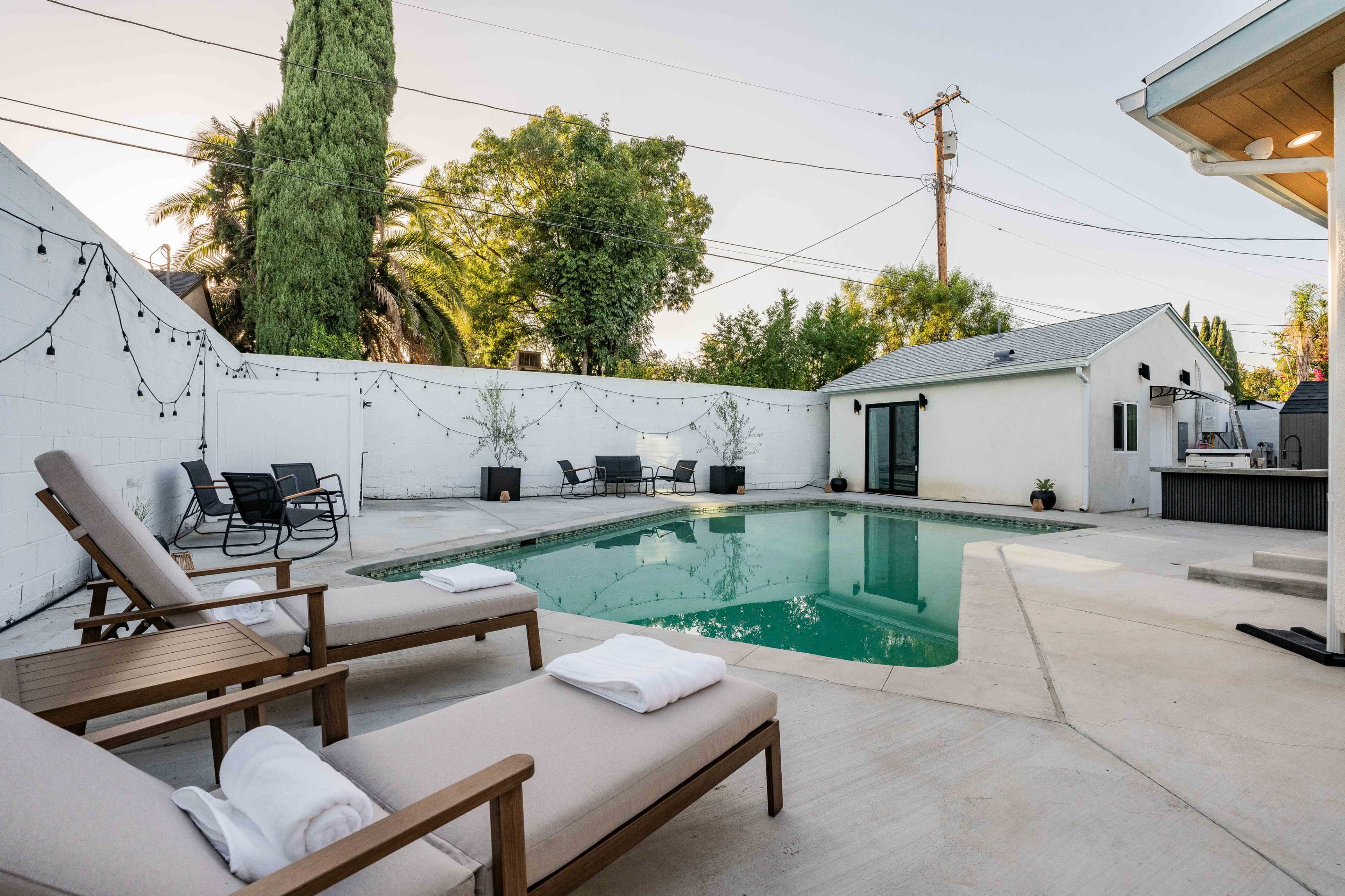 The image shows a pool area with lounge chairs and a landscaped backyard, enclosed by a white wall and trees.