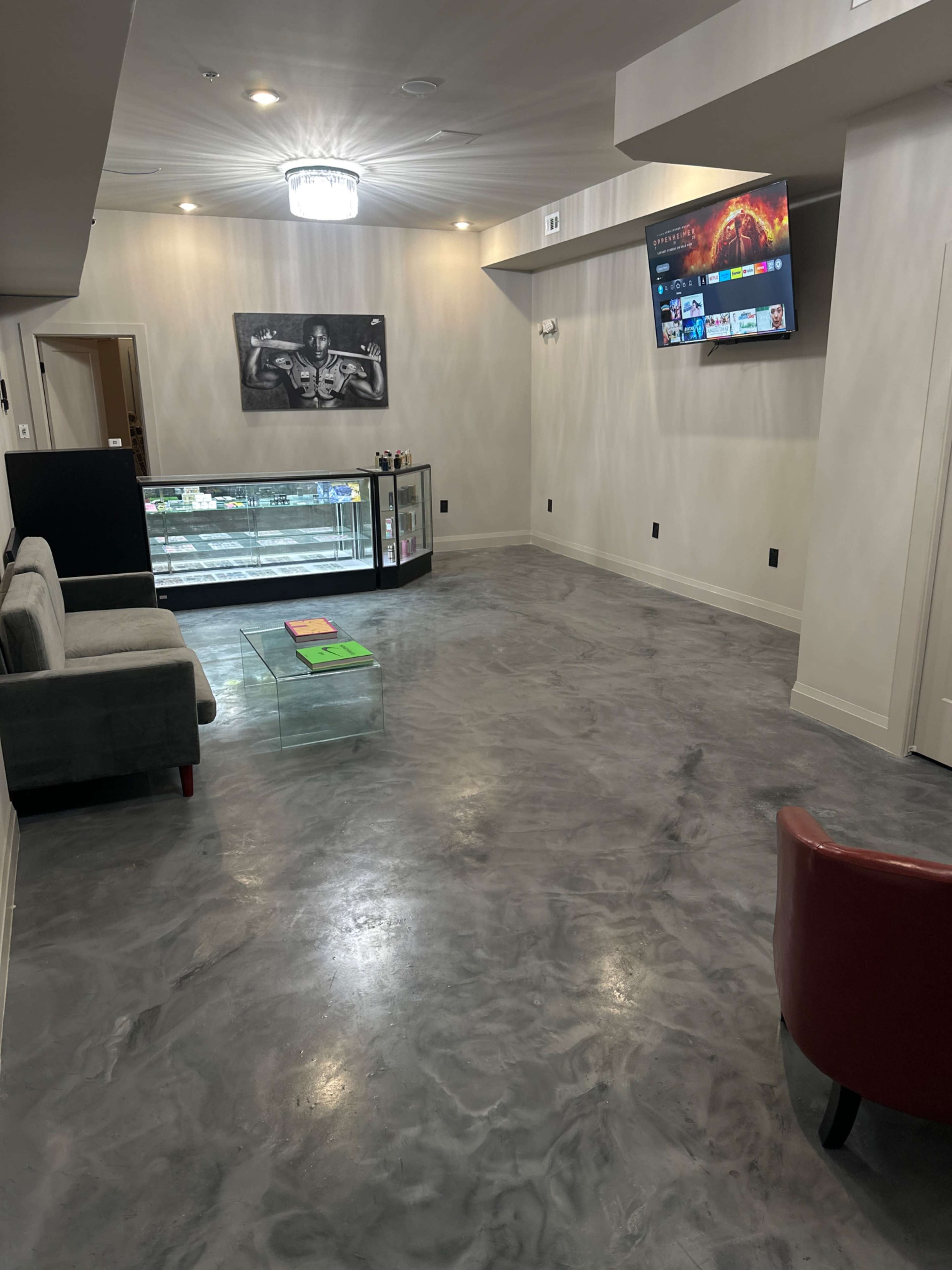The room features a gray polished concrete floor, a gray sofa, a red chair, a television on the wall, and a display cabinet.