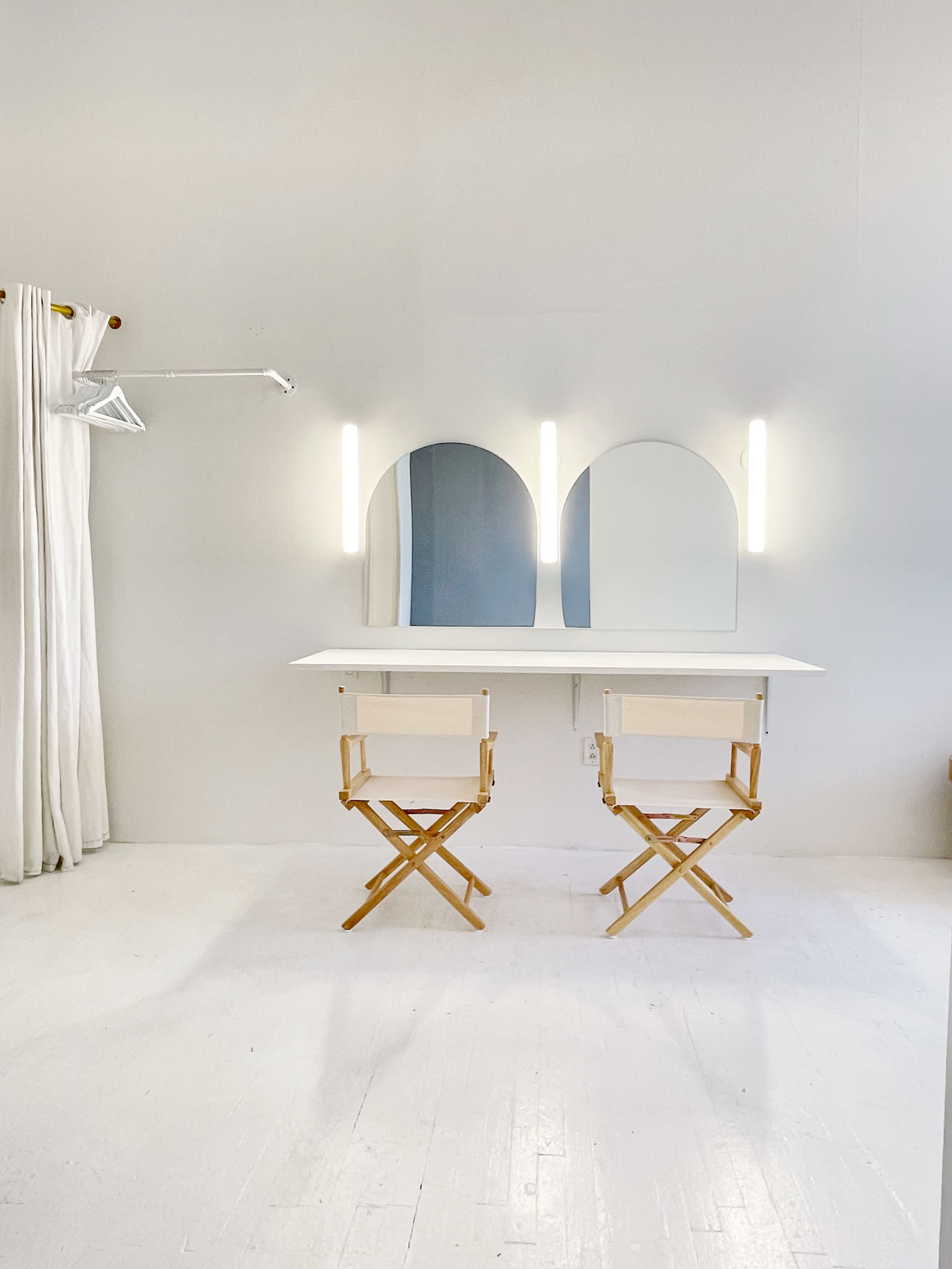 The image shows a minimalist dressing area with two wooden director's chairs facing a white table and three arched mirrors illuminated by vertical light fixtures.