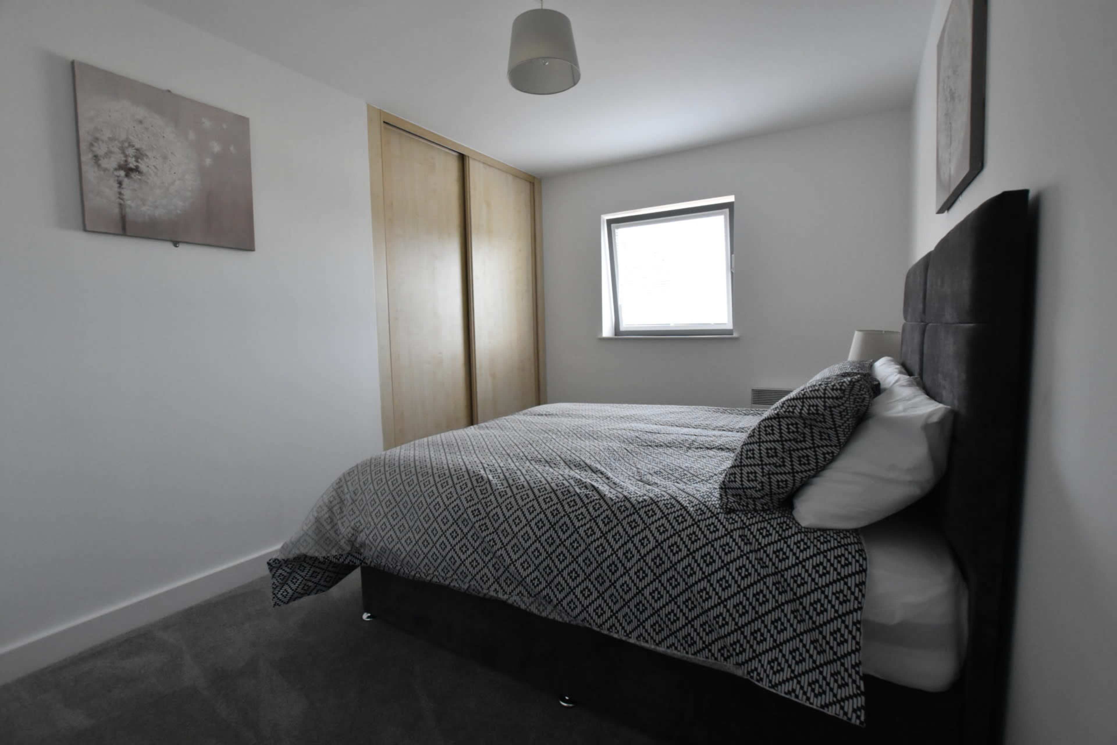 A neatly arranged bedroom featuring a bed with patterned bedding, a window with a gray frame, and a wooden wardrobe in the background.