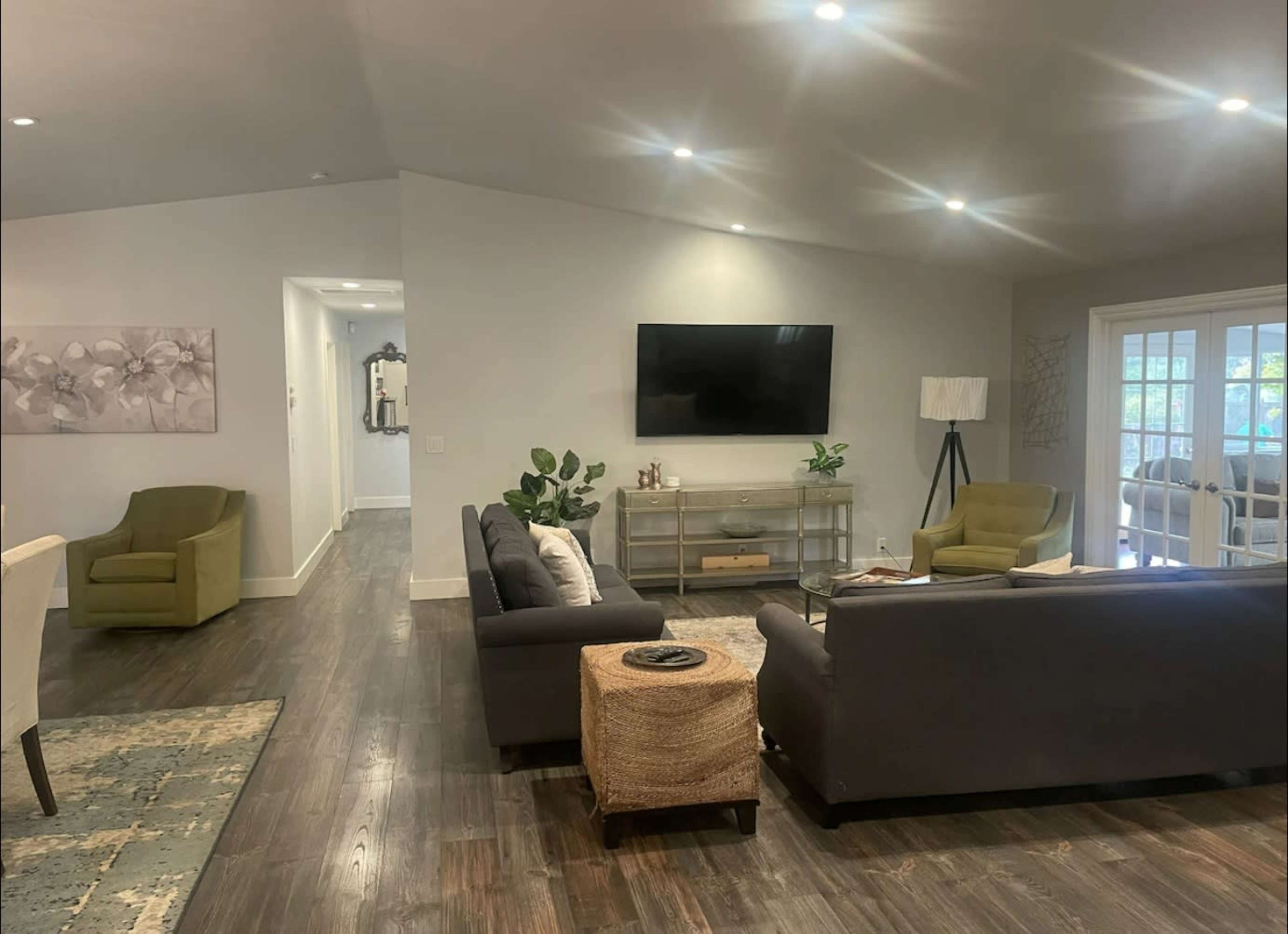 A modern living room with two green armchairs, a dark sofa, a glass coffee table, and a large television mounted on the wall.