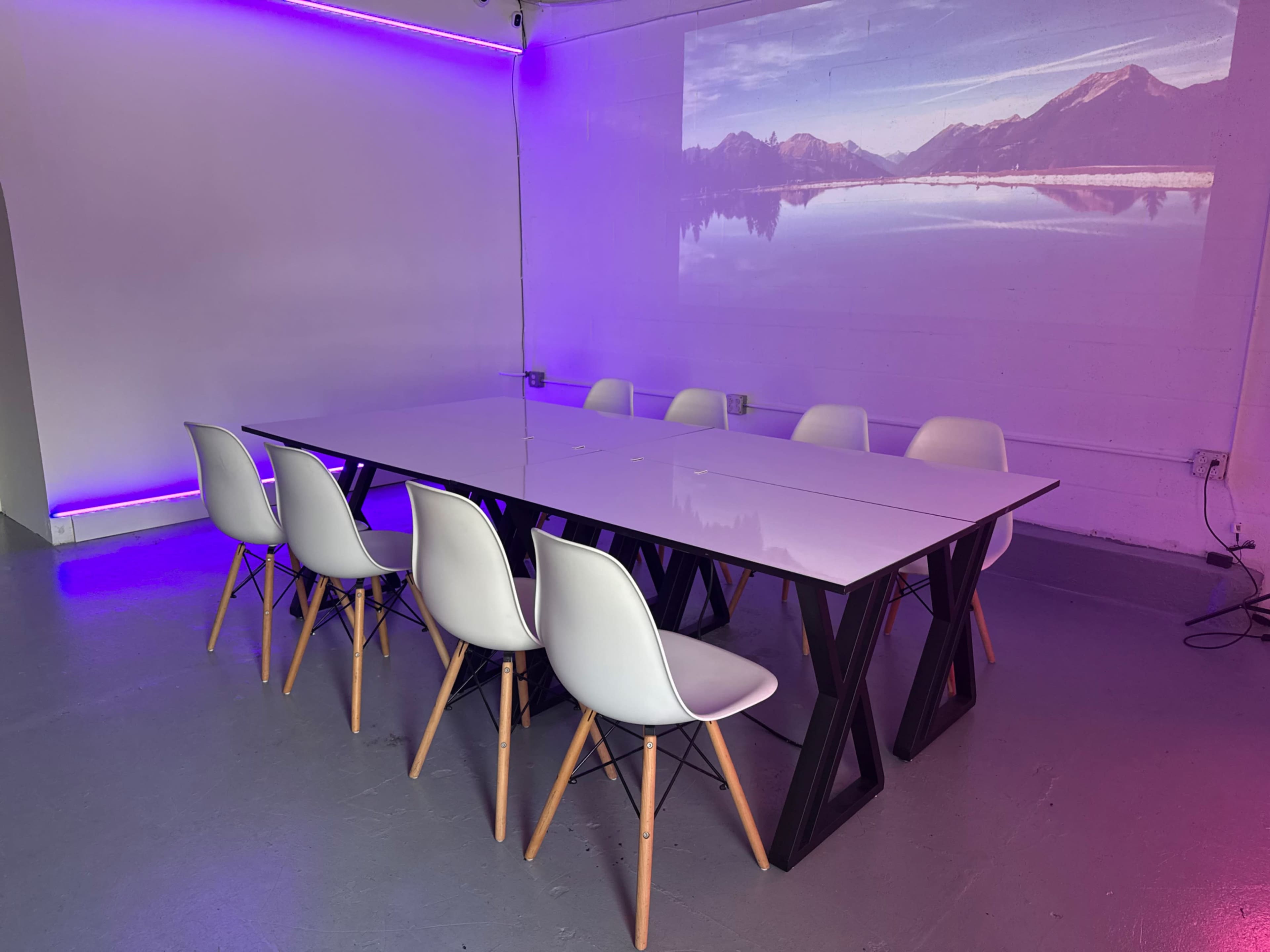 A modern meeting room features a long white table surrounded by white chairs, with a projection of a mountain landscape on the wall.