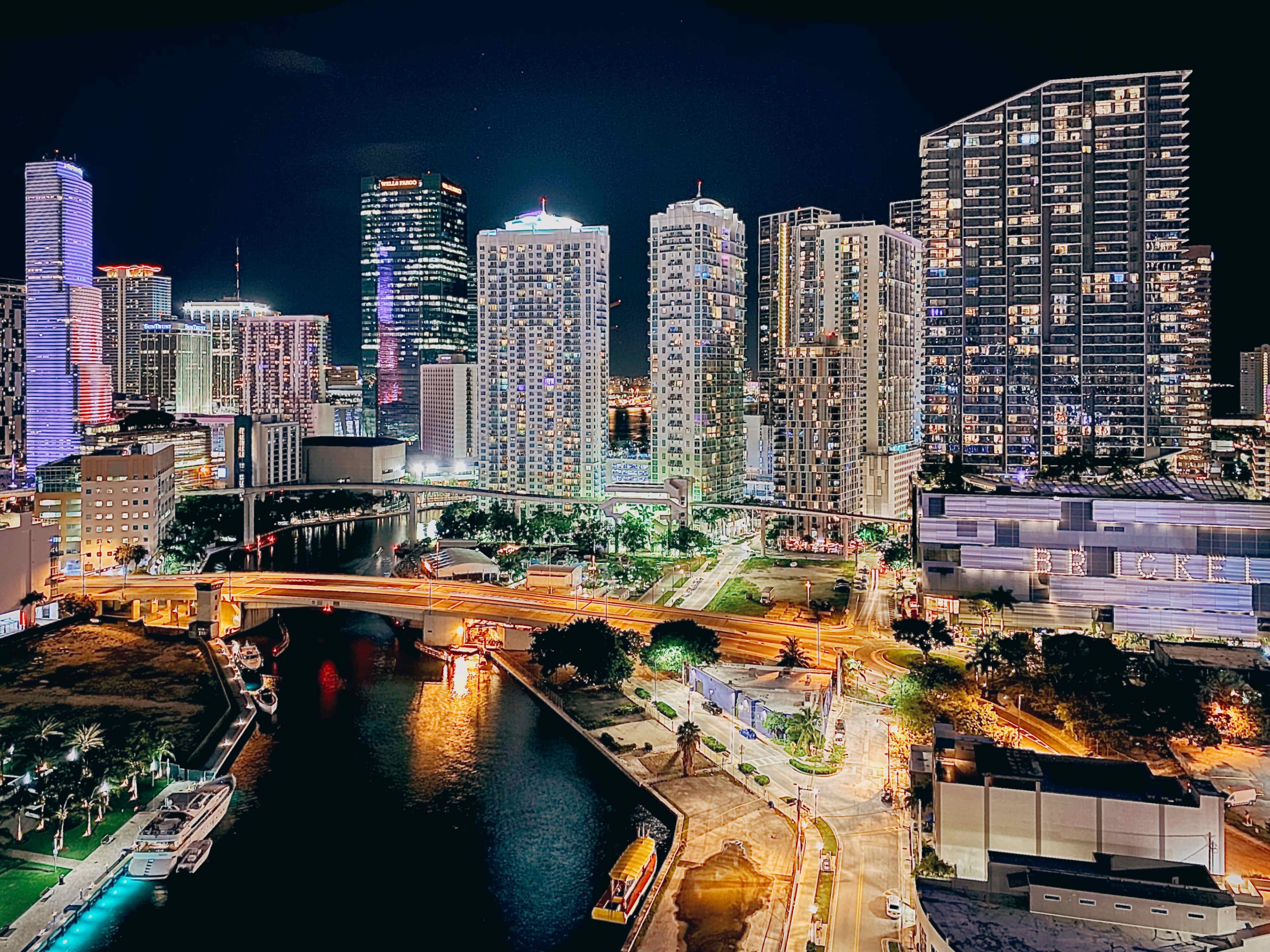 A vibrant city skyline at night, featuring tall buildings illuminated by colorful lights beside a winding river with a bridge and parked boats.