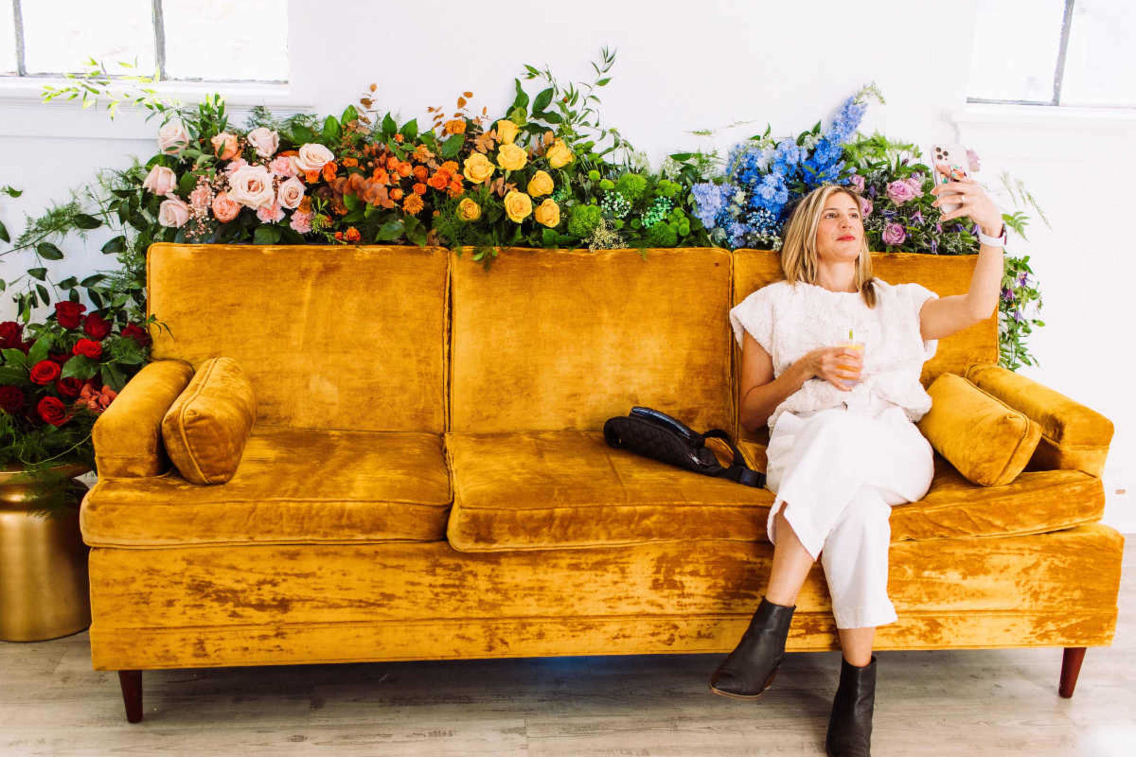 A woman is seated on a yellow sofa surrounded by a vibrant arrangement of flowers.