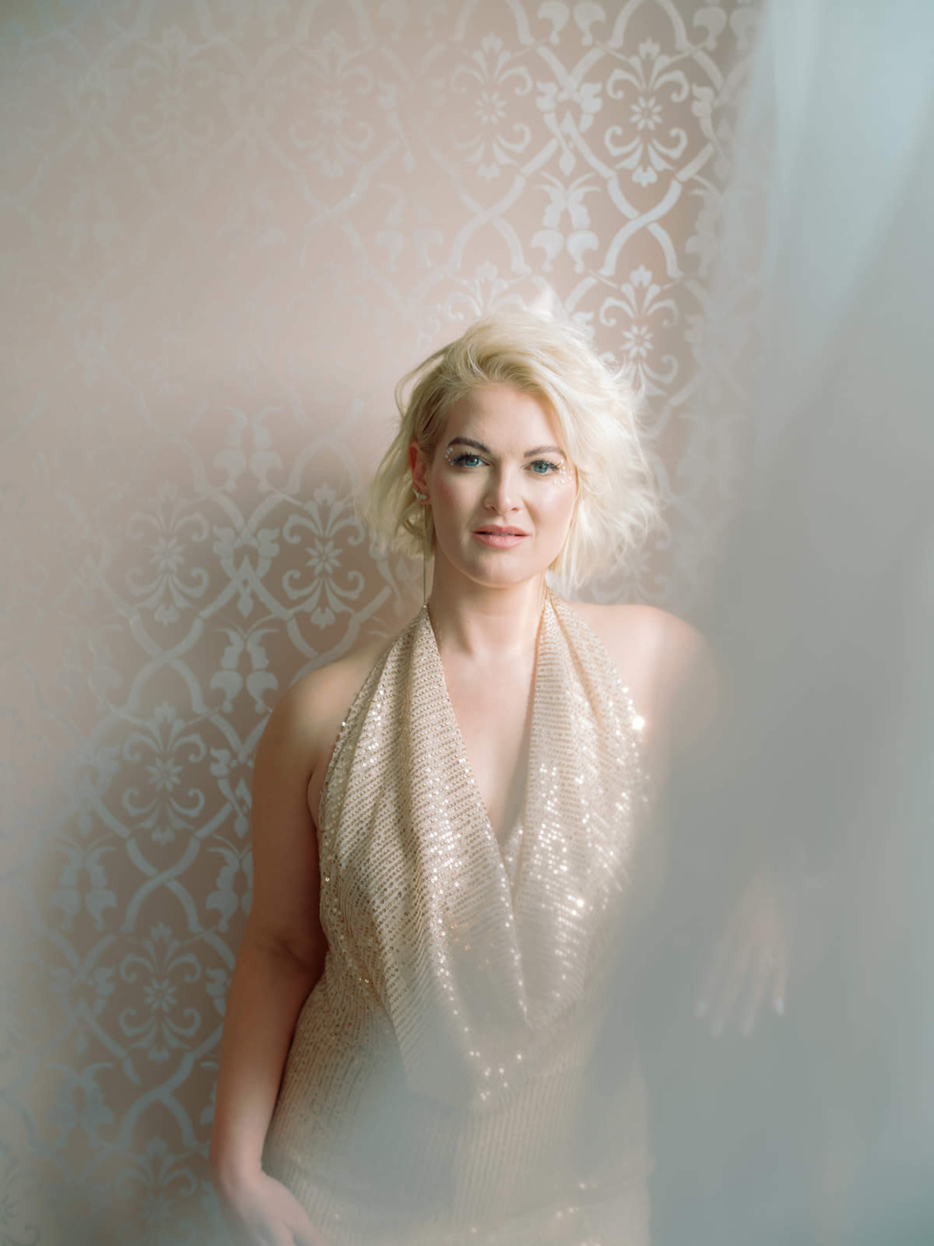 A woman wearing a sequined gold dress poses in front of a patterned wall, partially obscured by a sheer curtain.