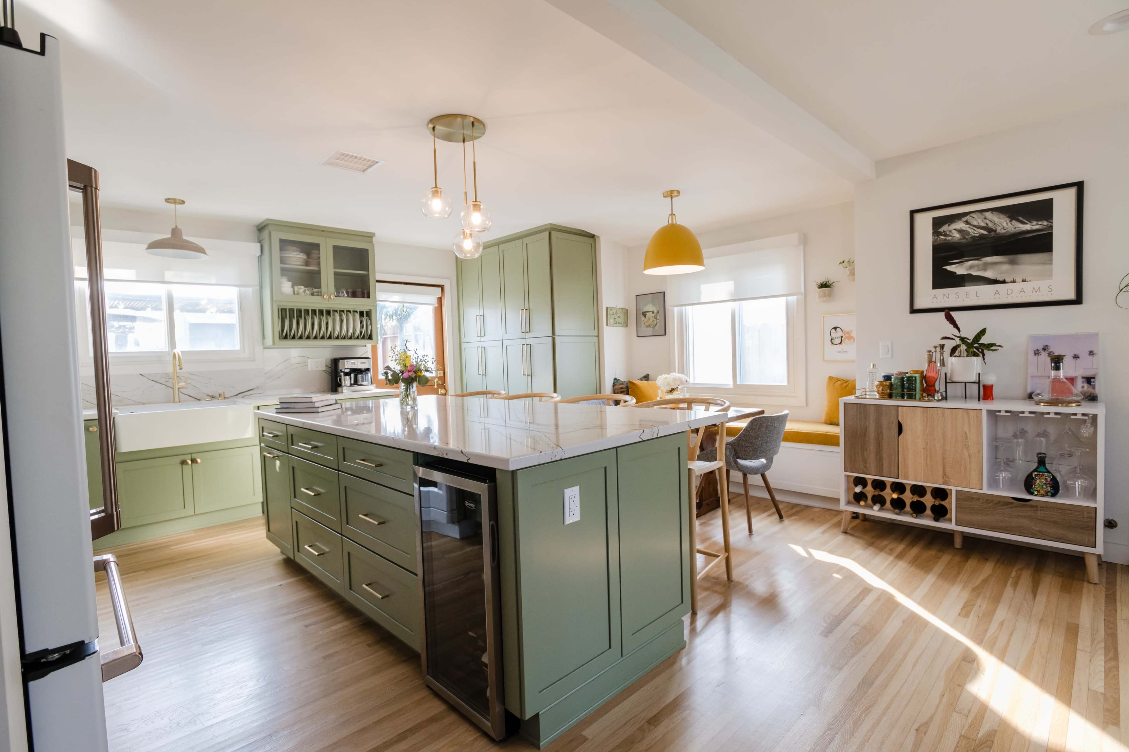 A modern kitchen featuring an island with green cabinetry, a marble countertop, and a dining area with a wooden table and yellow pendant light.