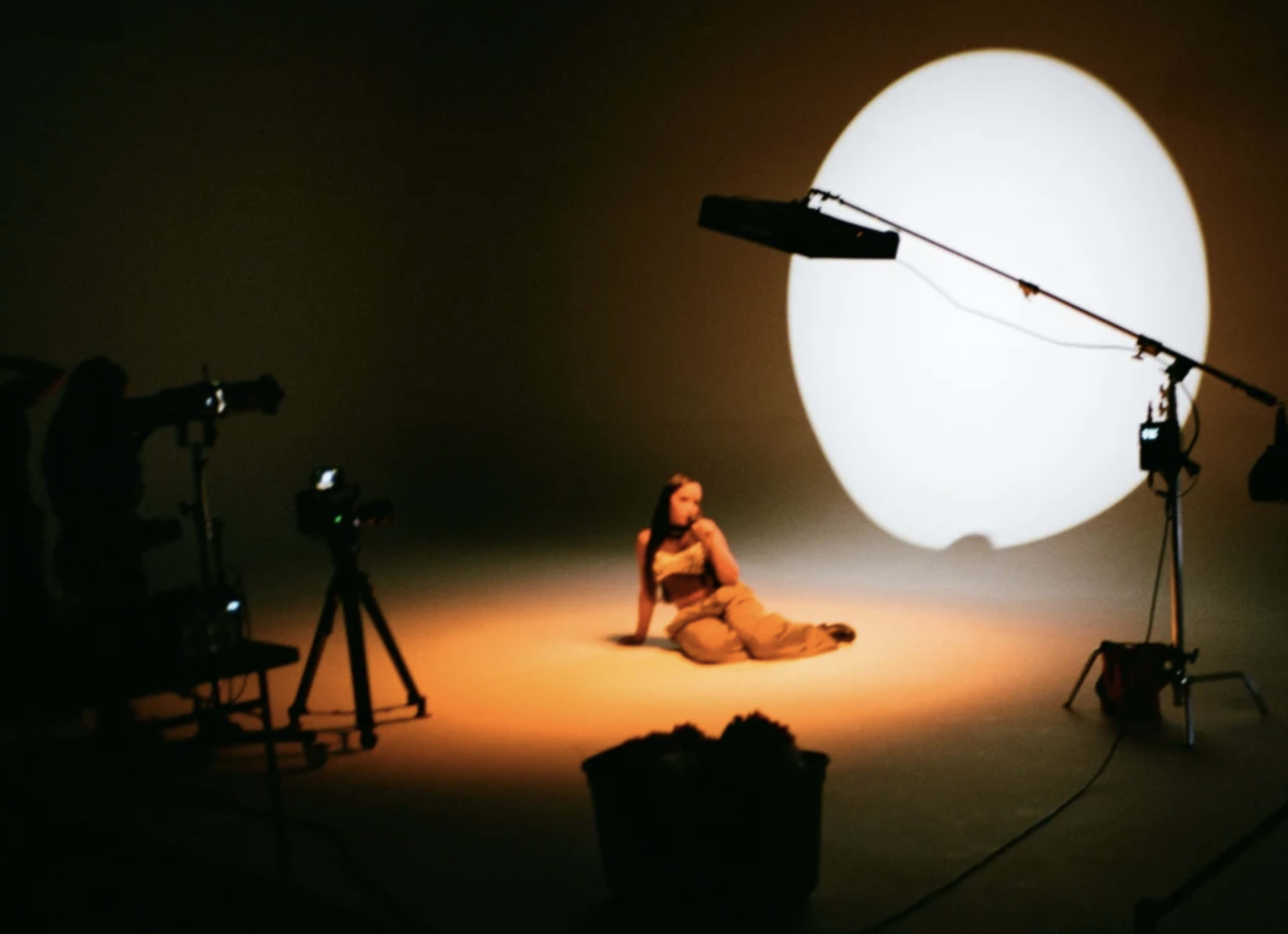 A person sits on the floor of a studio, illuminated by a large circular backdrop light, with cameras positioned around the scene.