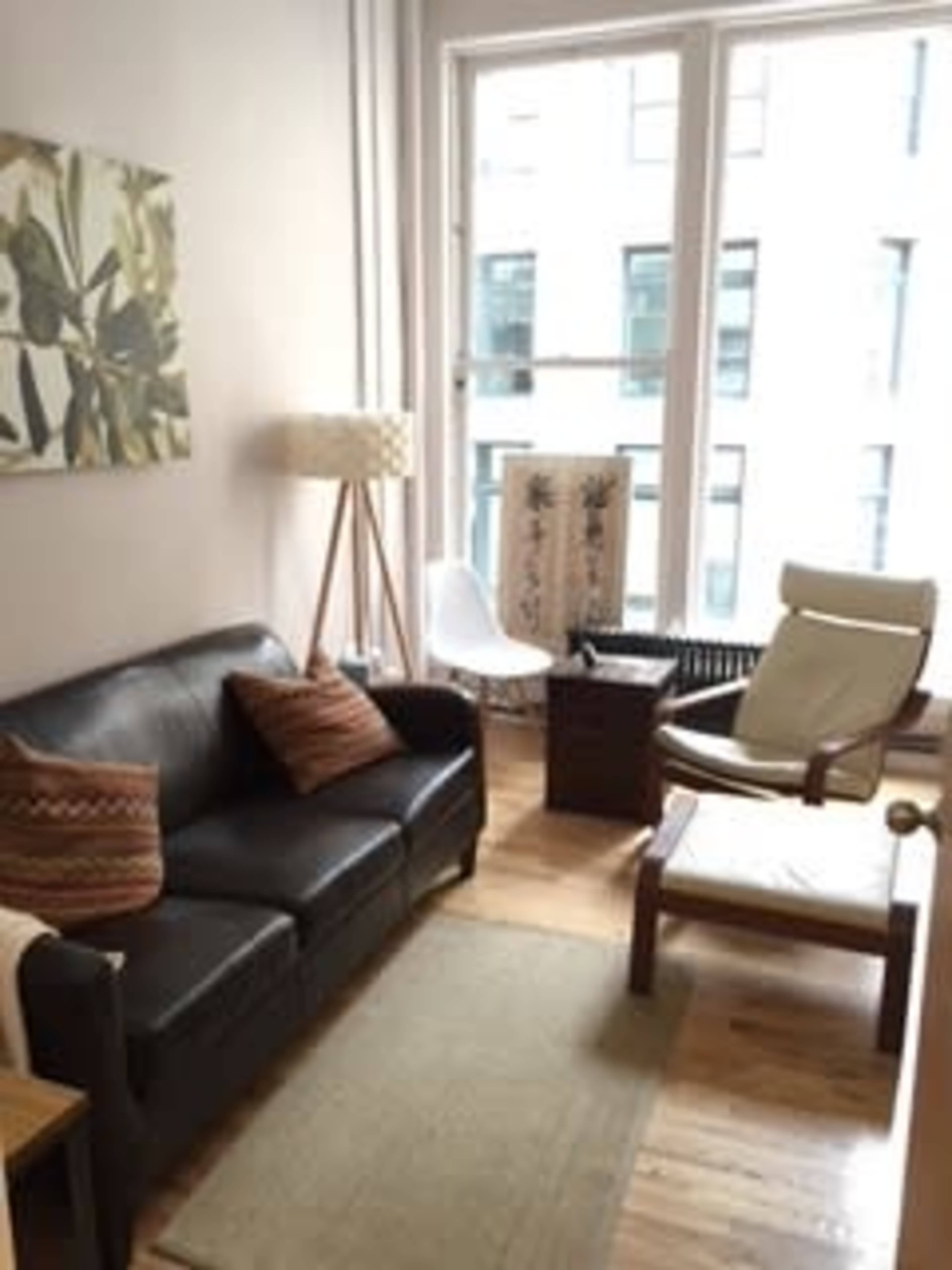 A small living room with a leather couch, a floor lamp, a white chair, and a wooden table next to a large window.