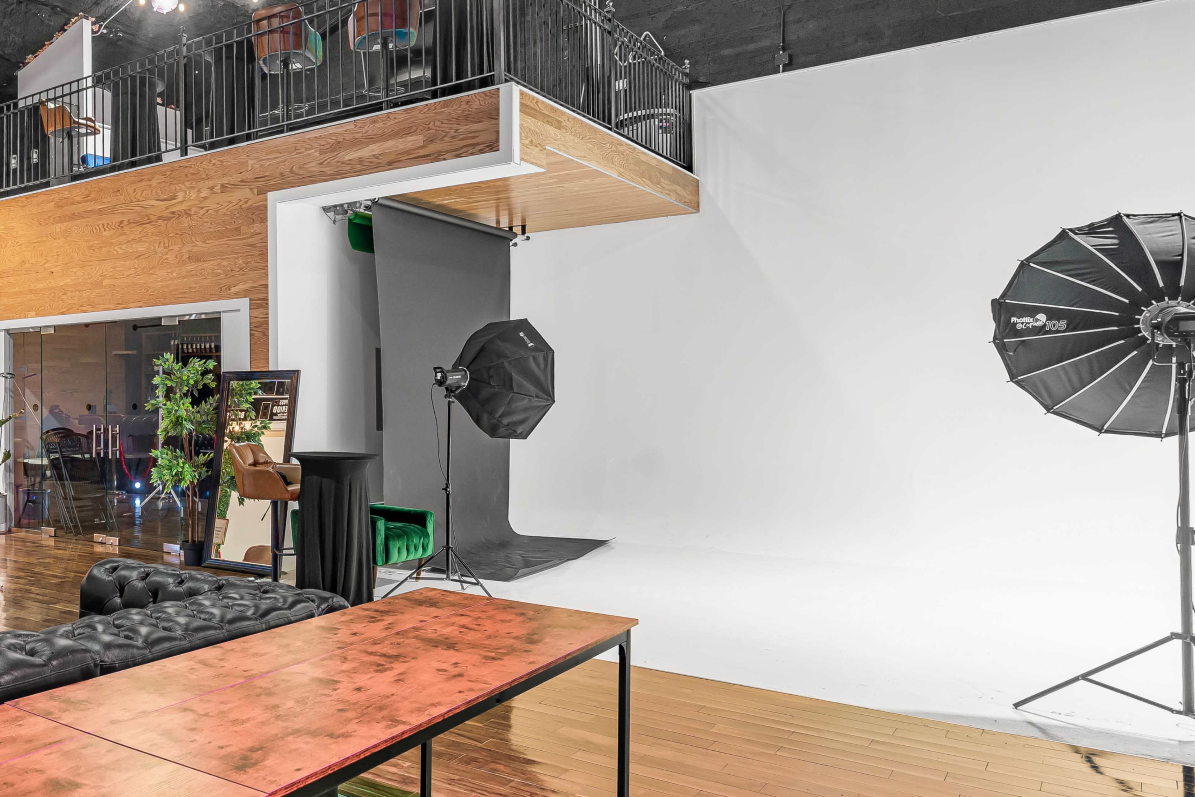 The image shows a photography studio with a large white backdrop, two softboxes, and a seating area featuring a black couch and a wooden table.