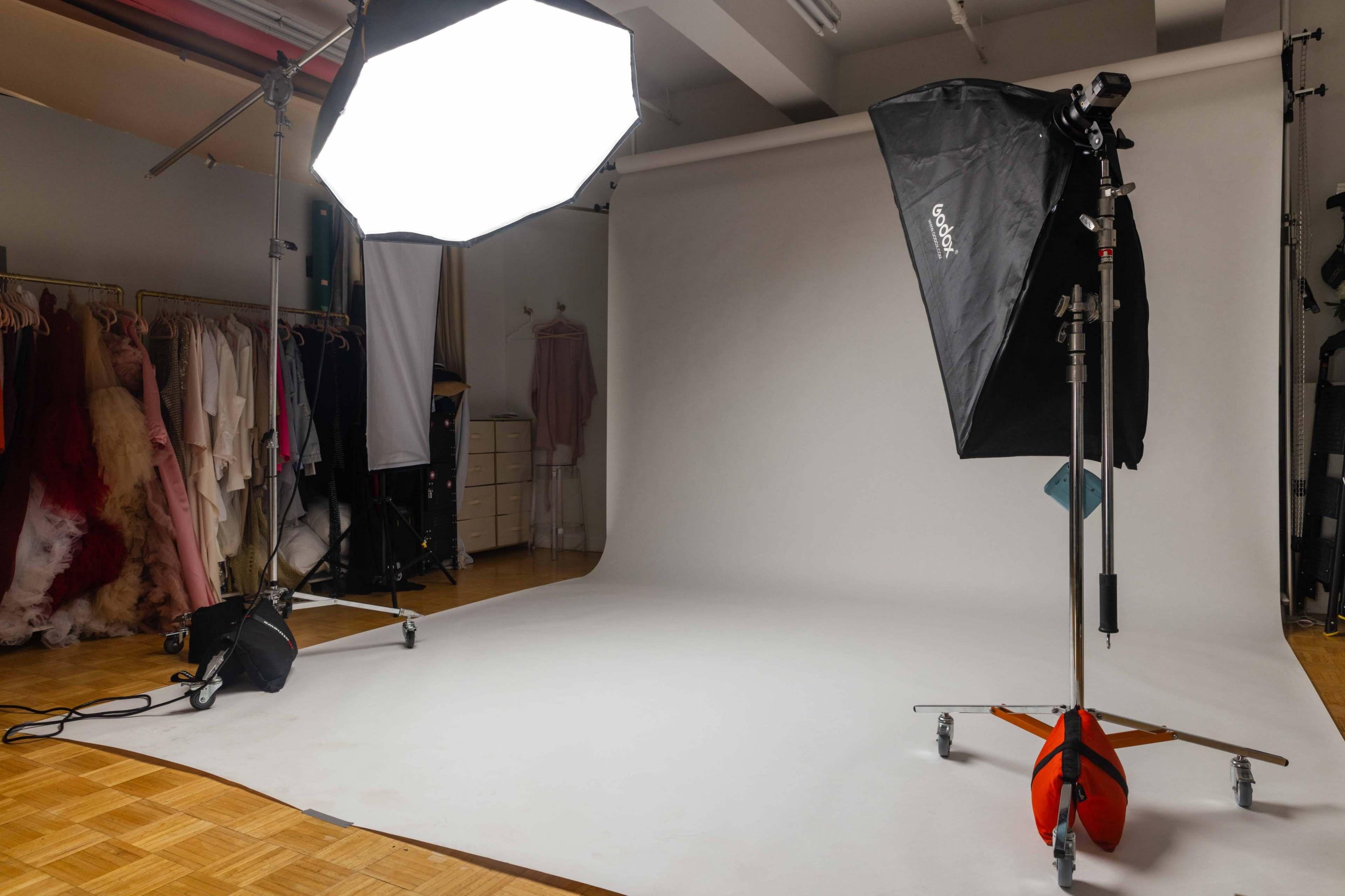 A photography studio setup with a large softbox light, a black umbrella reflector, and a smooth white backdrop, surrounded by hanging garments.