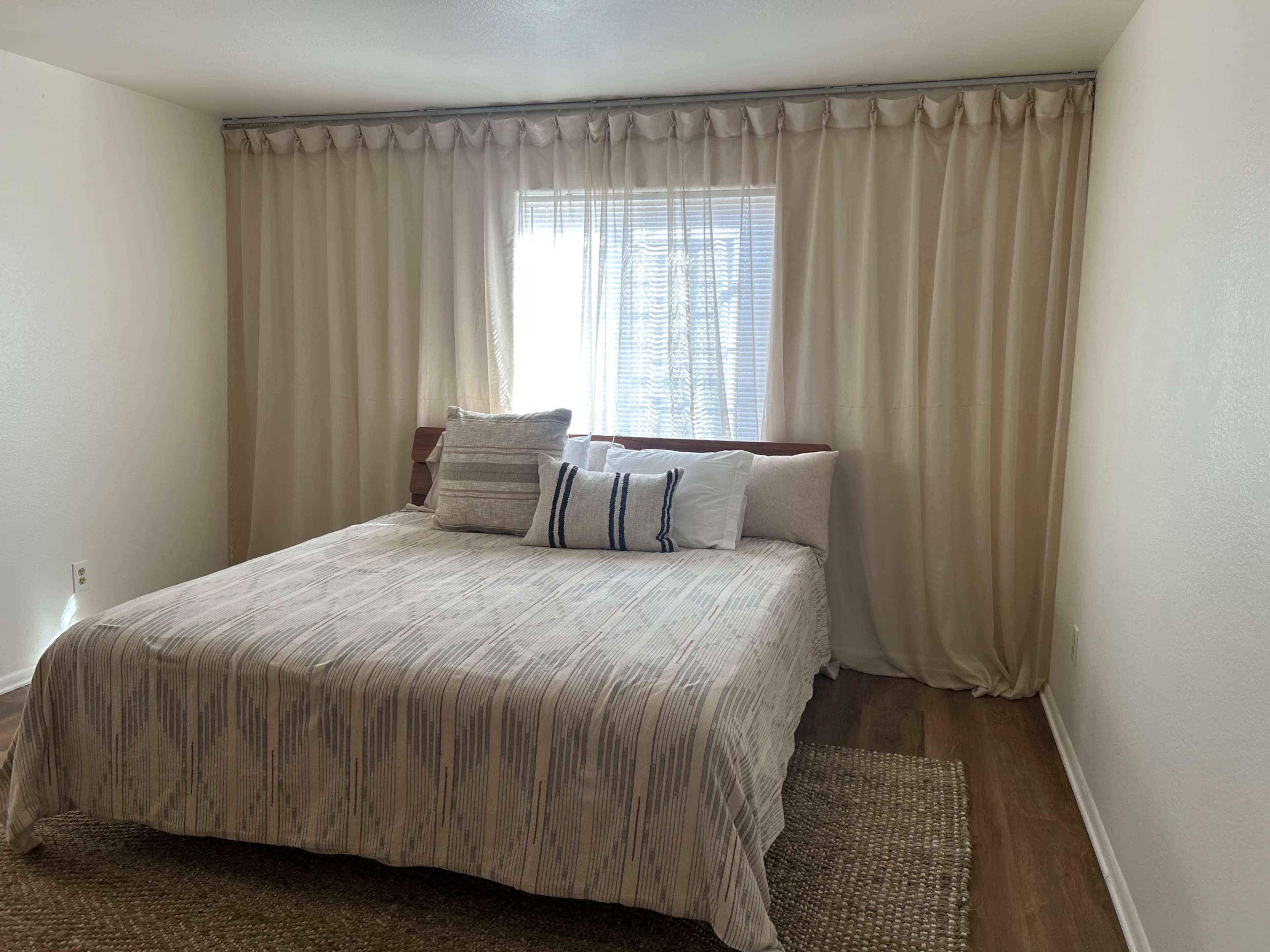 The image shows a simple bedroom with a large bed covered in a patterned blanket, white pillows, and a window partially covered by beige curtains.