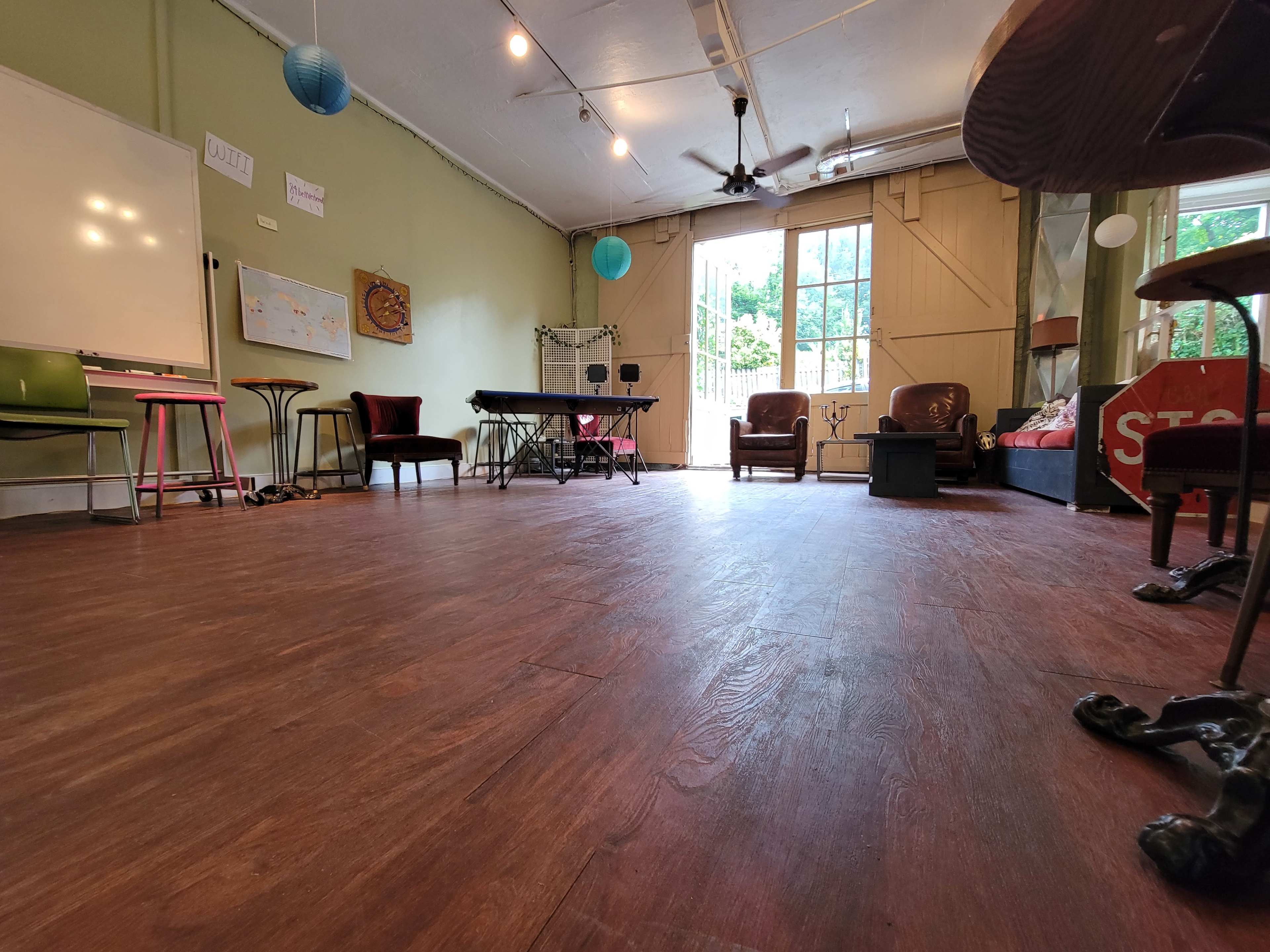 The image shows a spacious room with hardwood flooring, a few chairs, a table, and large windows allowing natural light to enter.