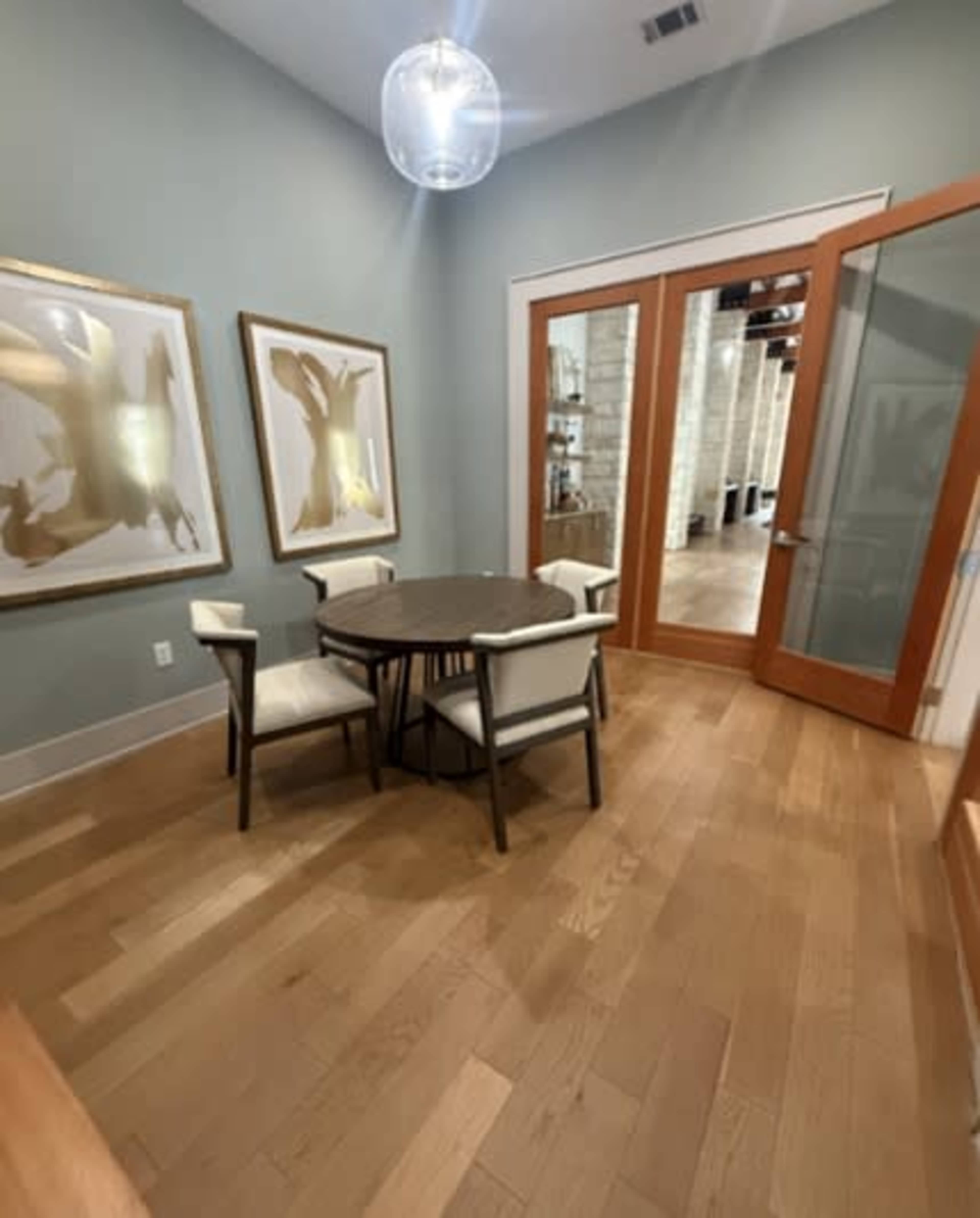 The image shows a small dining area featuring a round table surrounded by four chairs, with two framed artworks on the wall and double glass doors leading to another room.