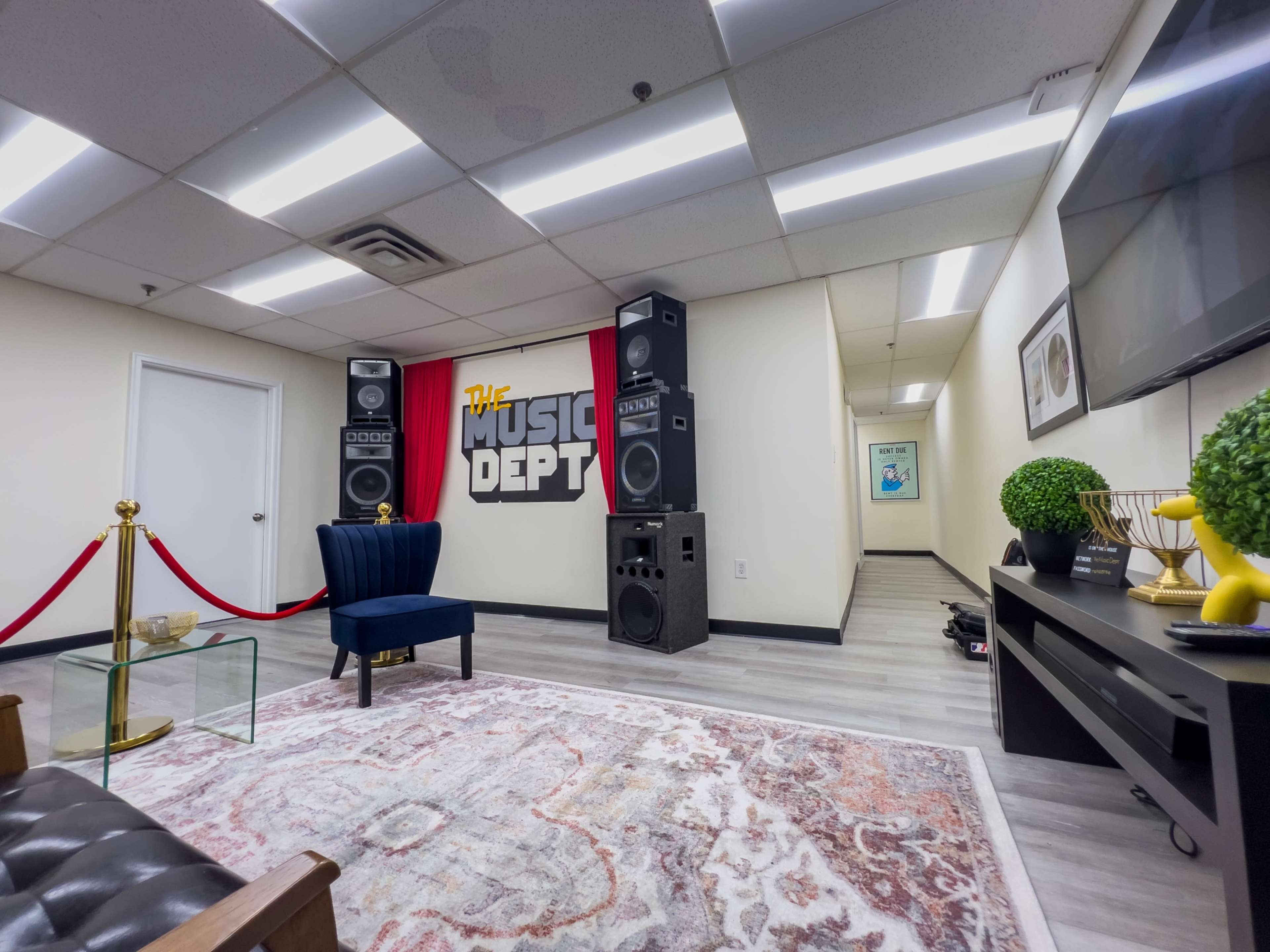 The room features a seating area with a blue chair, two large speakers, a red backdrop displaying "The Music Dept," and a hallway leading to another space.