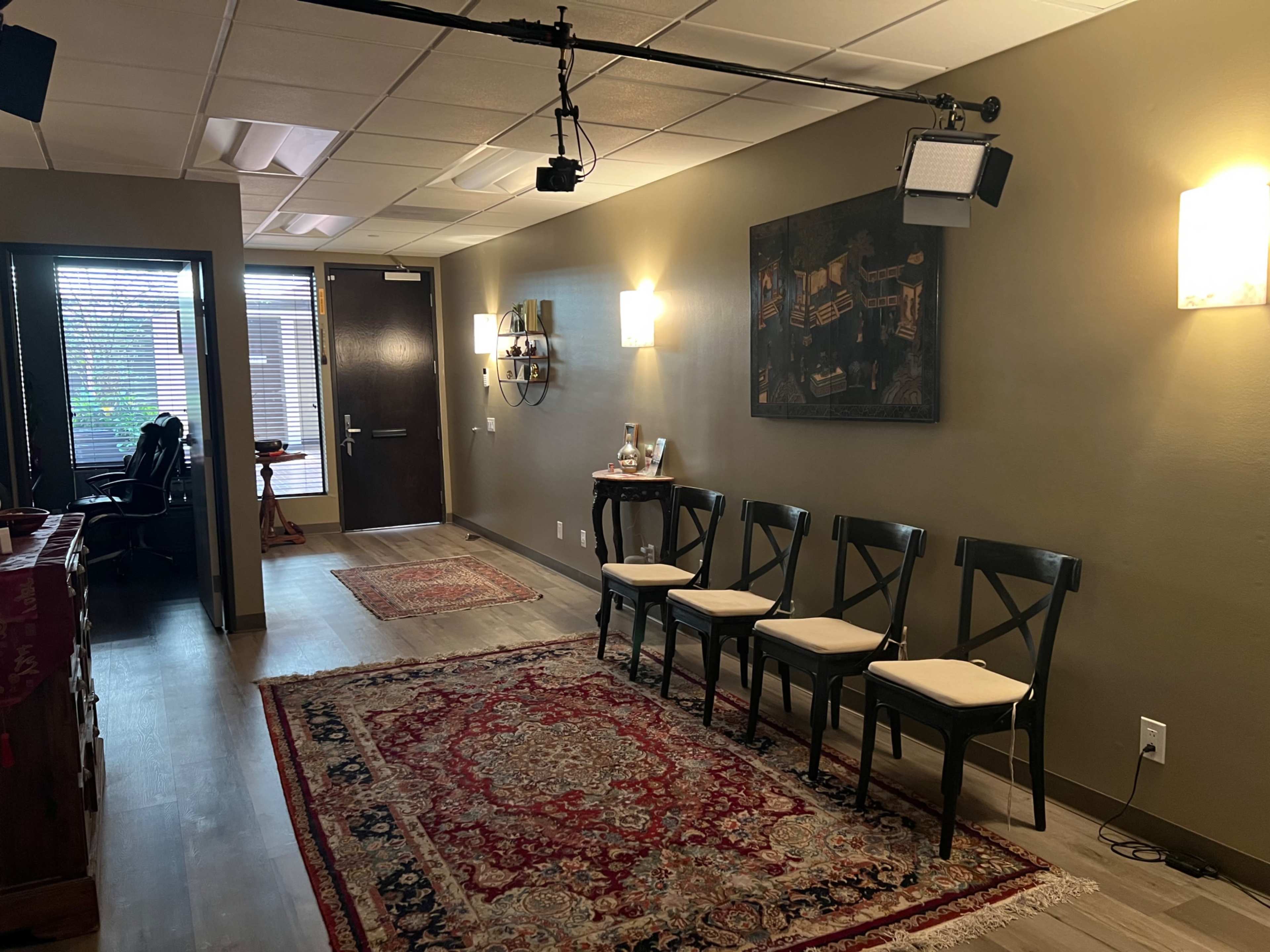 The image shows a waiting area with four black chairs aligned against a wall, a decorative rug on the floor, and artwork hanging above, leading to a doorway in the background.