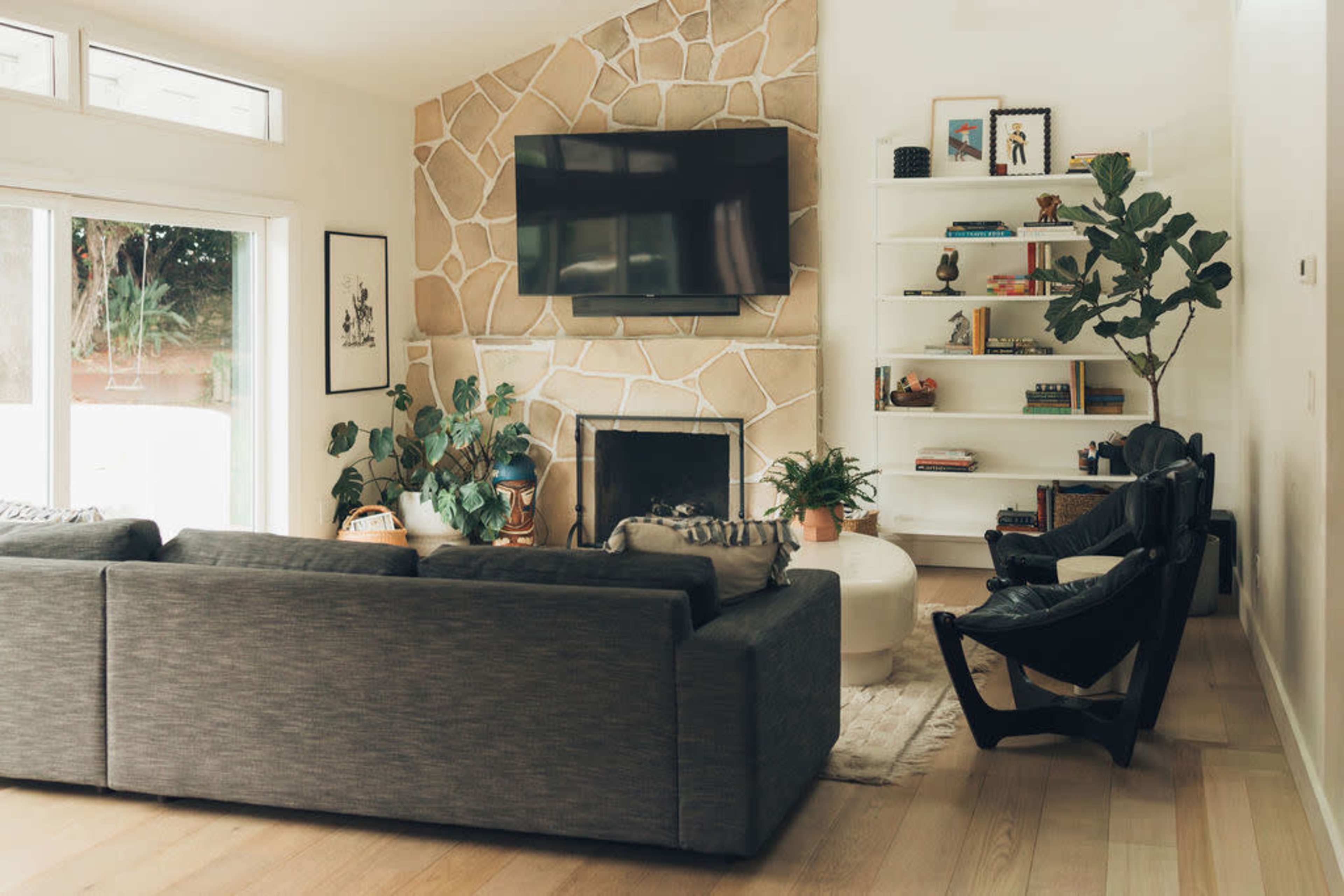 A modern living room features a stone fireplace, a television mounted above it, a large gray sectional sofa, and a bookshelf filled with decorative items and plants.