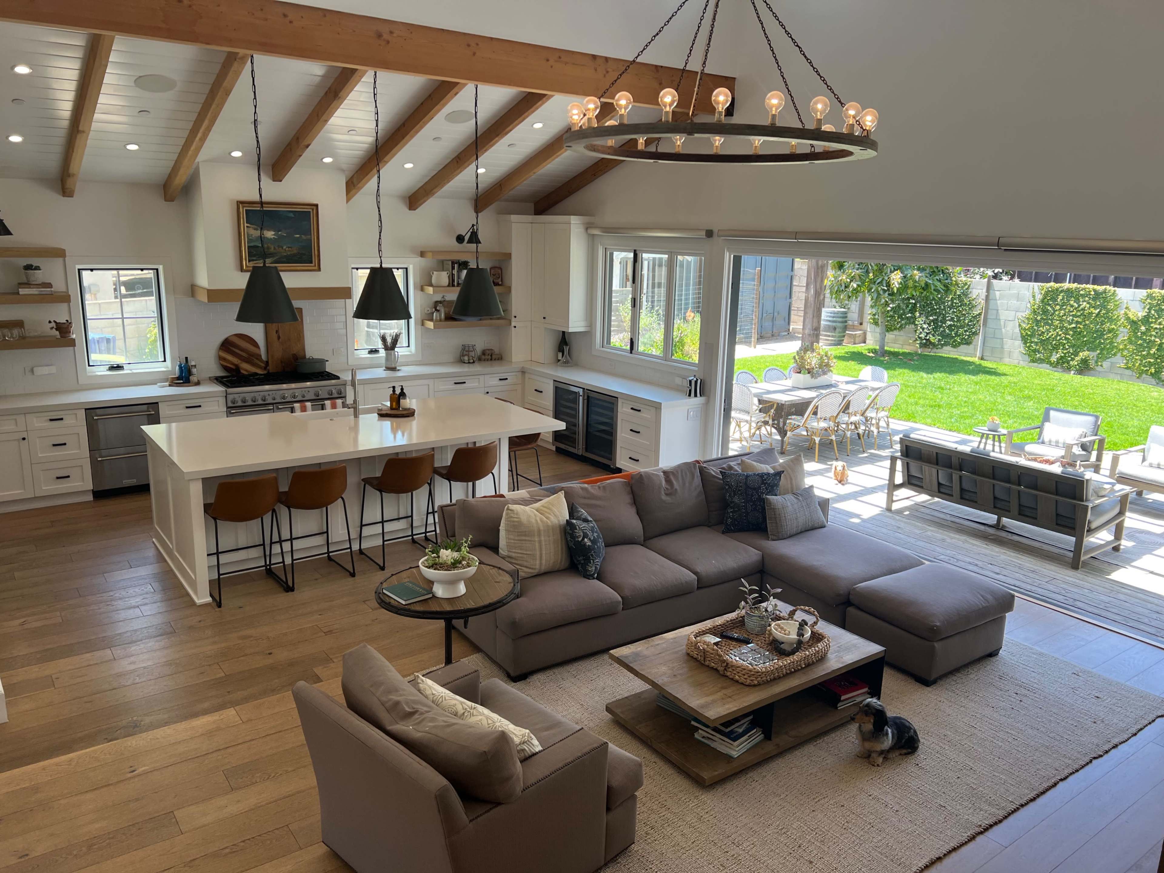 A spacious, modern open-concept living area featuring a kitchen with a large island, a comfortable sectional sofa, and a view of an outdoor dining space with a lush green lawn.
