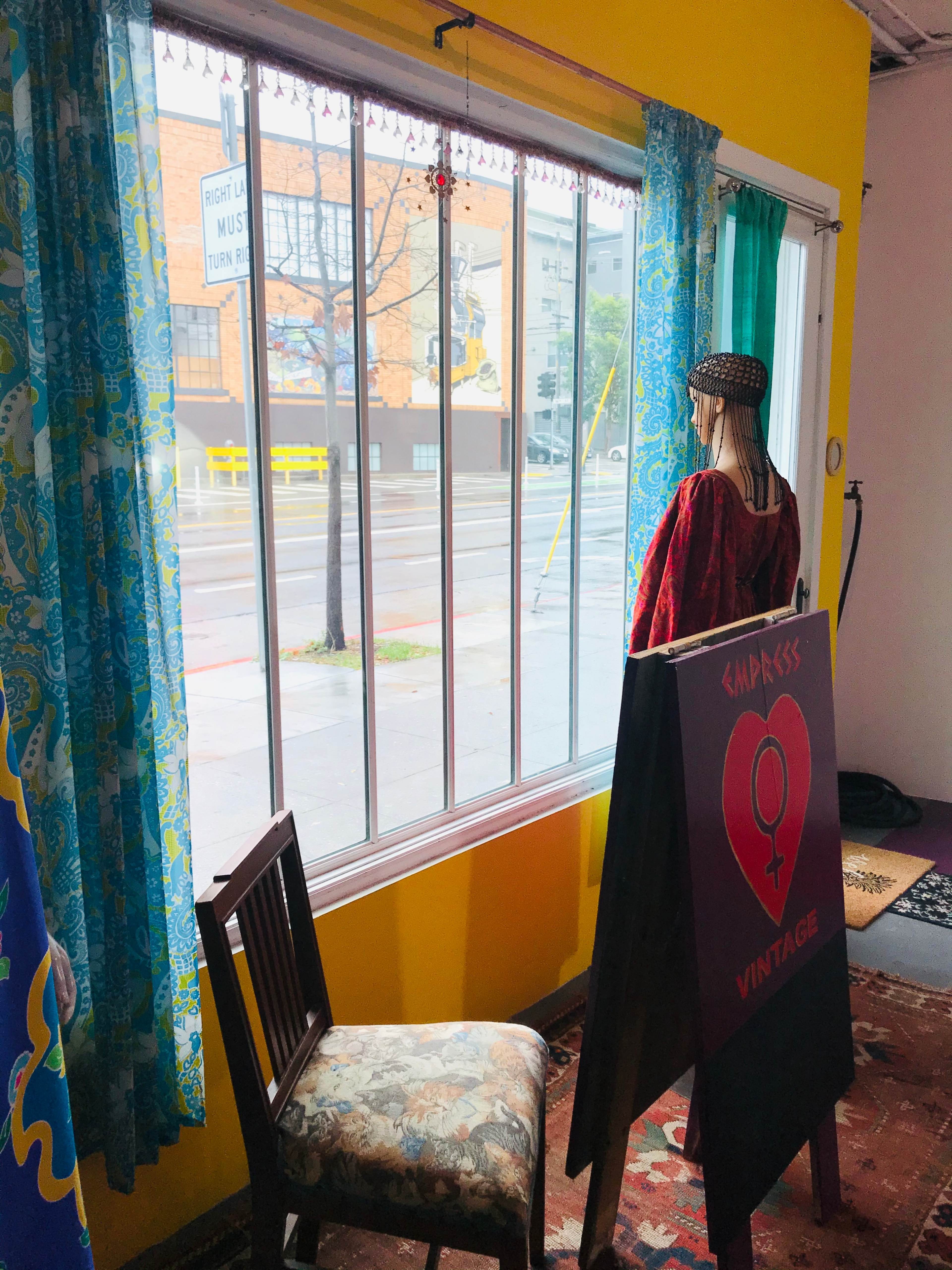 A mannequin dressed in vintage clothing stands by a large window, overlooking a rainy street with colorful buildings in the background.