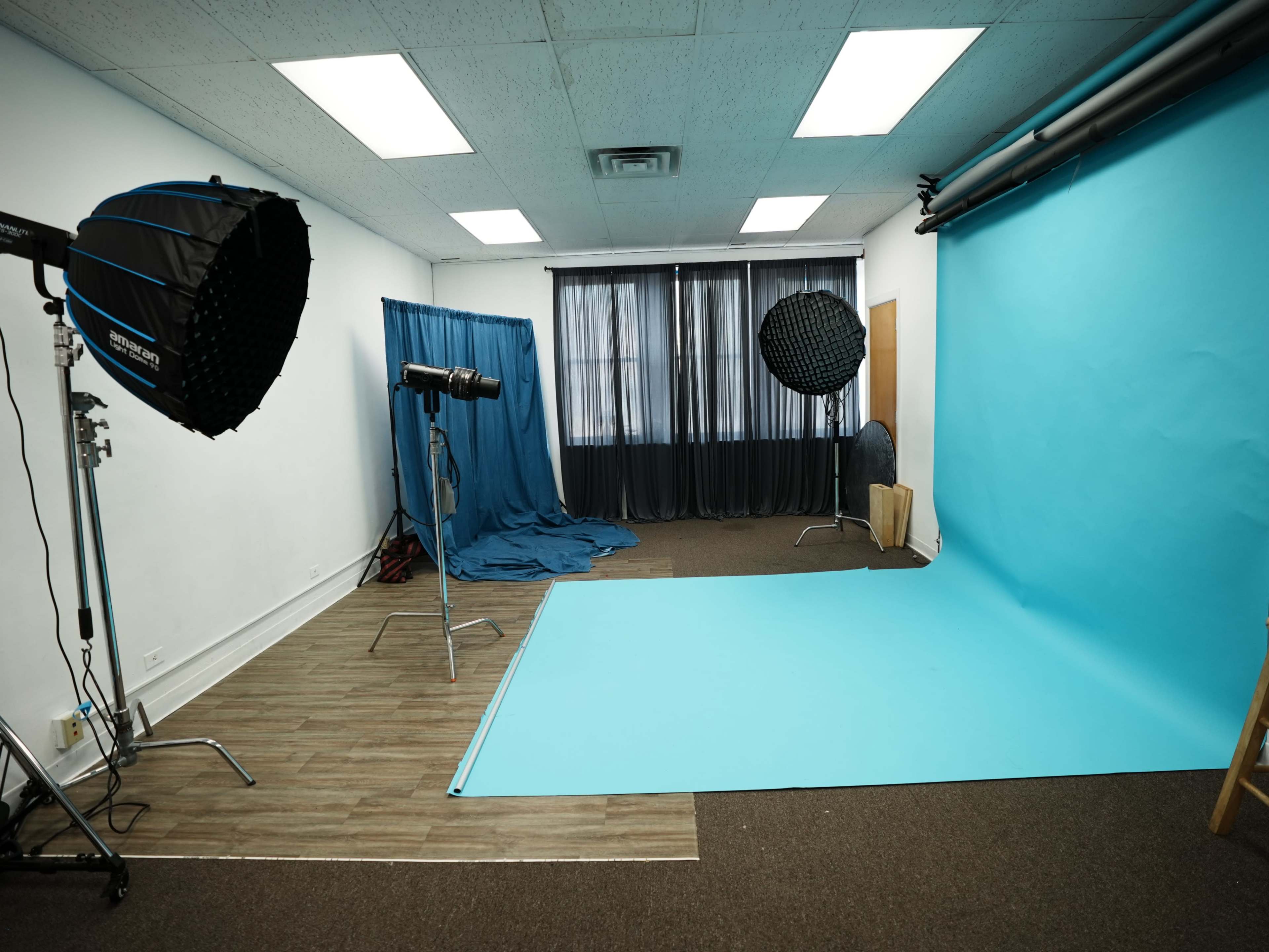 The image shows a photography studio with a blue backdrop, softbox lights, and a large window with curtains.