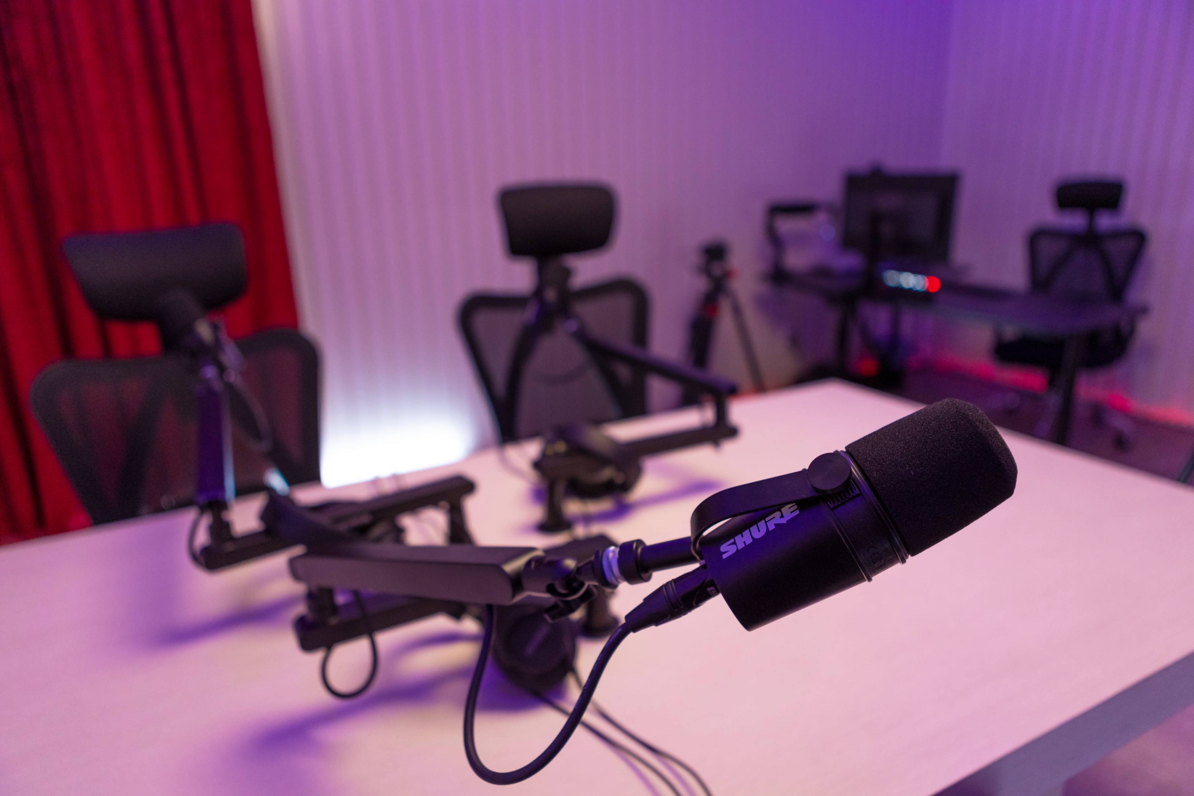 A podcast recording studio with multiple microphone setups on a table and seating in the background.