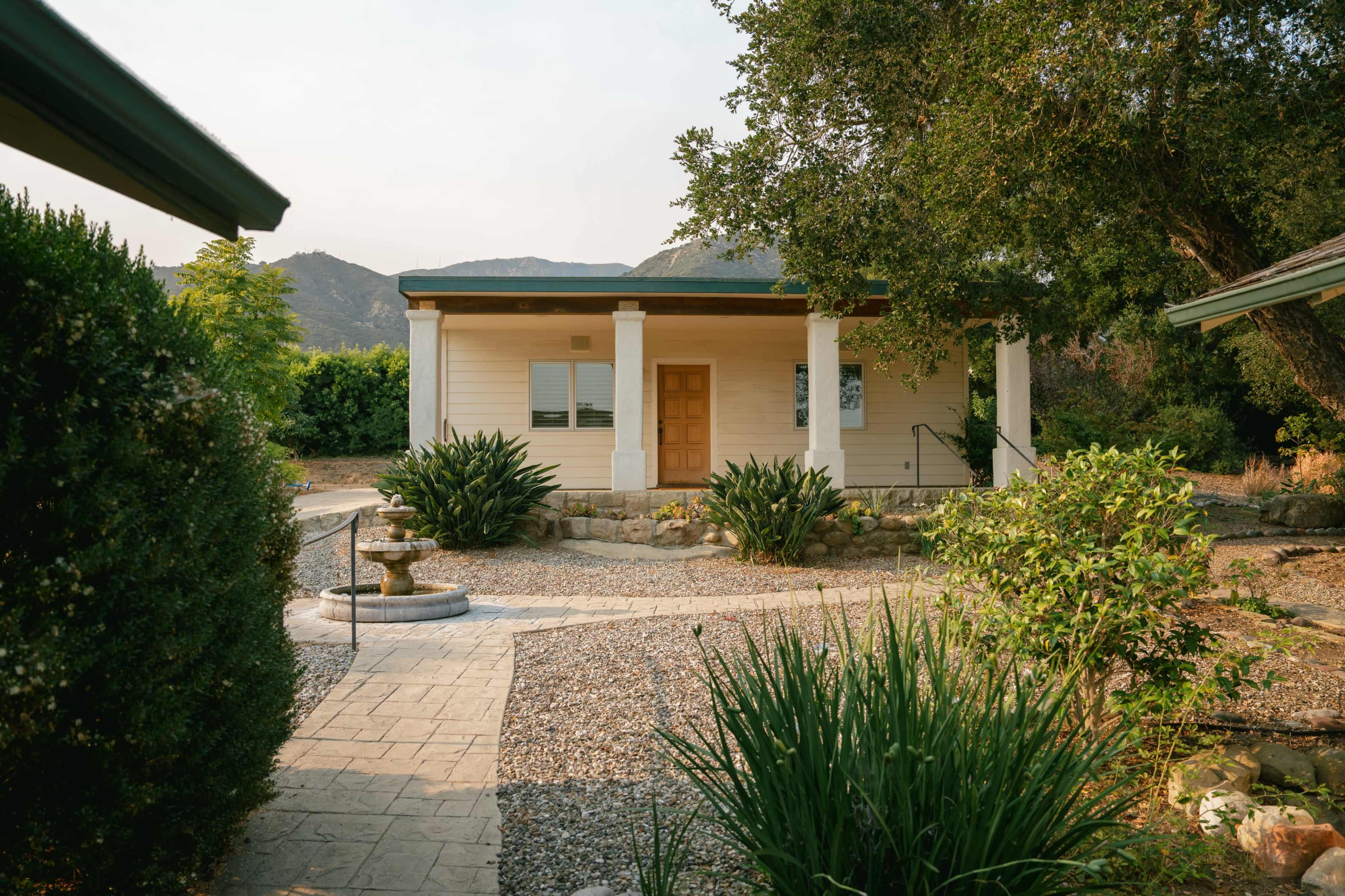 The image features a small house with a wooden front door, surrounded by landscaped gravel paths and greenery, set against a backdrop of mountains.