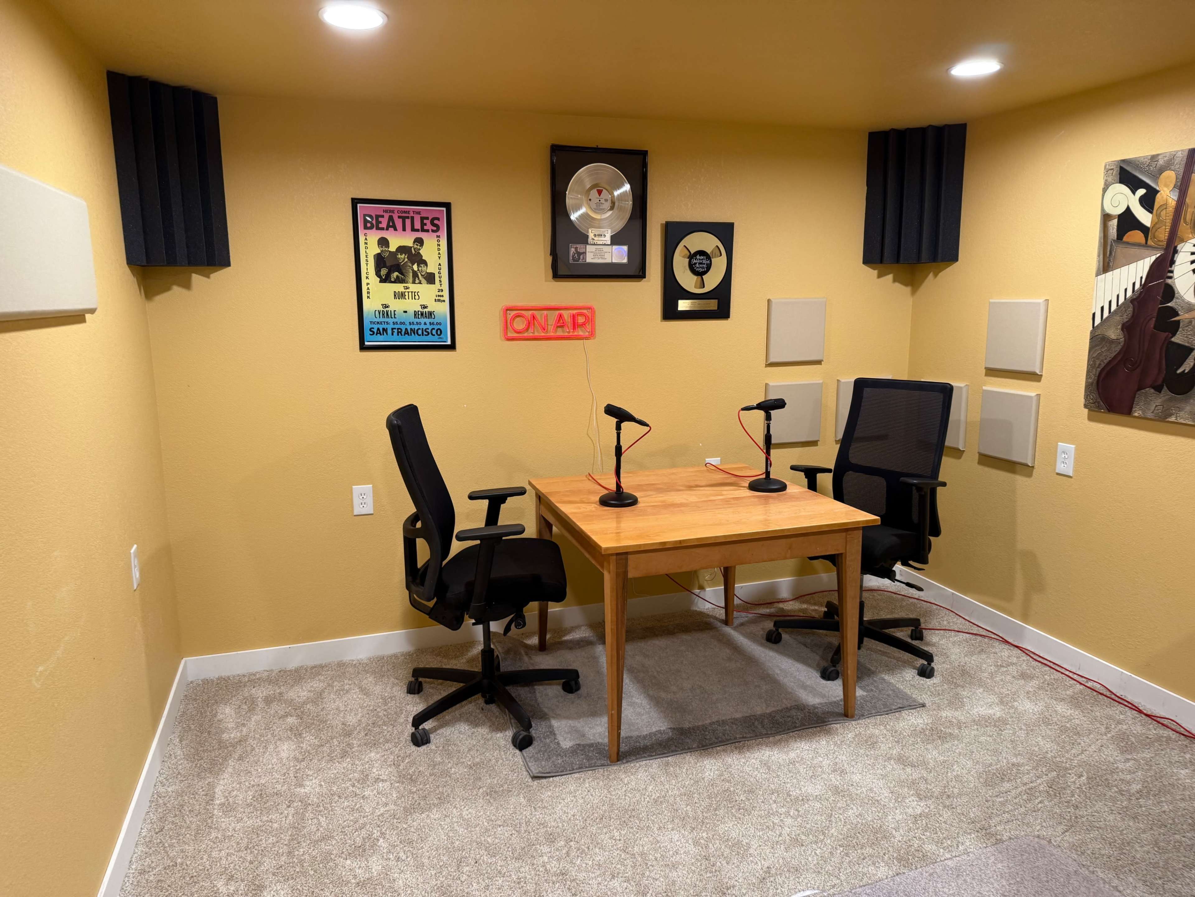 The image shows a podcast recording studio with a wooden table and two black chairs, framed music memorabilia on the walls, and soundproofing panels.