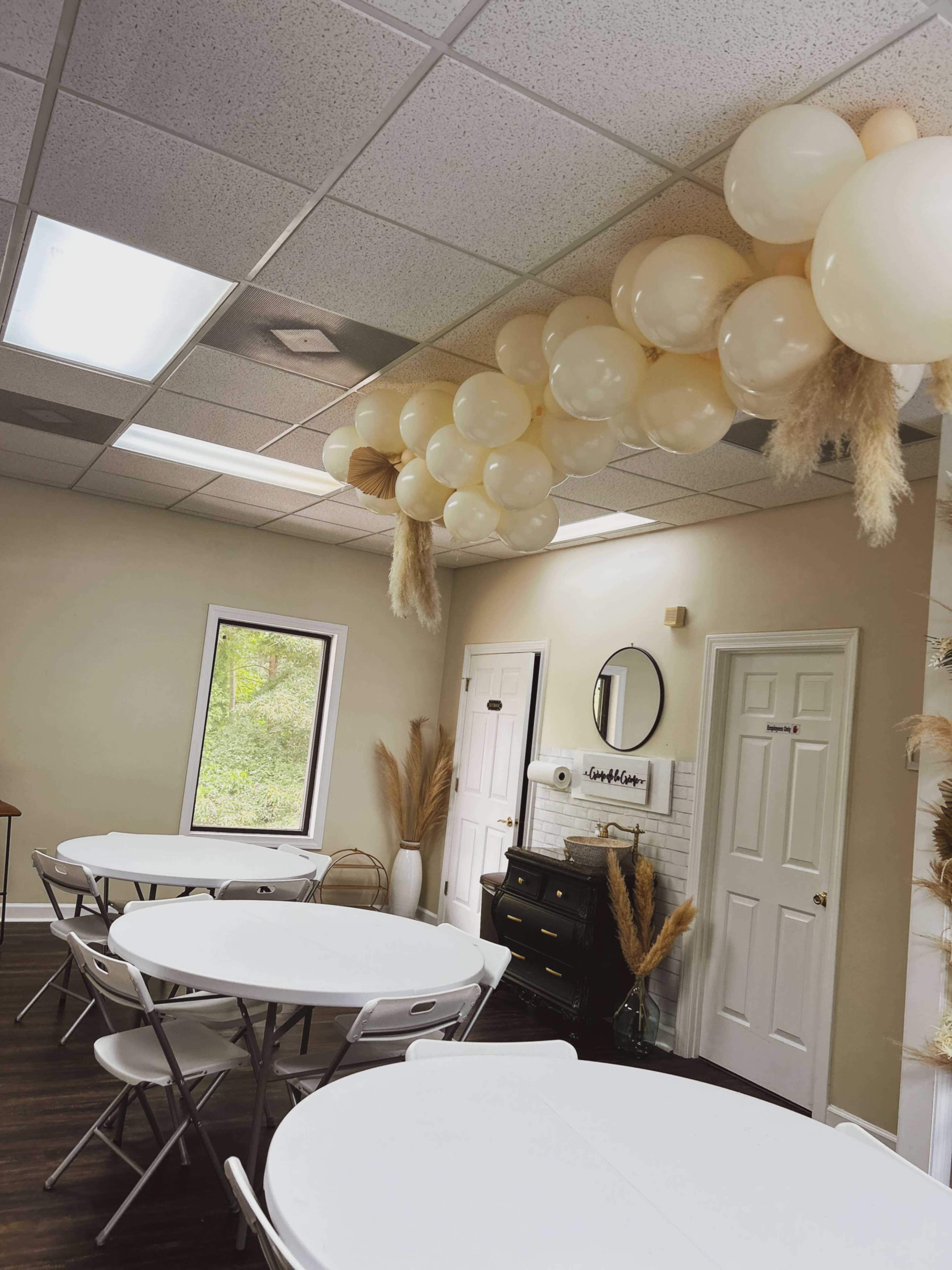 A series of beige balloons is arranged overhead in a room with white tables and chairs, featuring a window and a decorative wall.