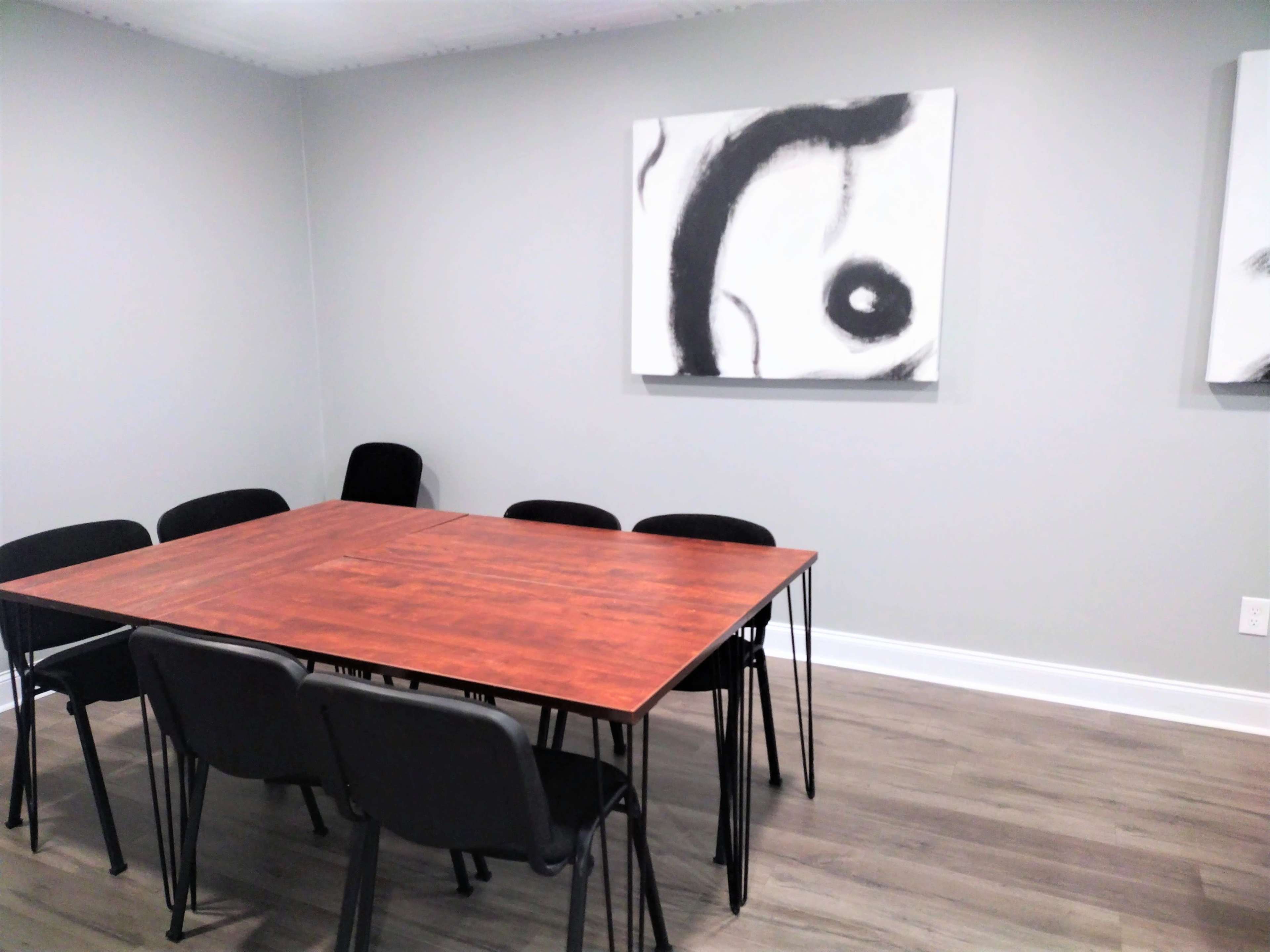 A square wooden table surrounded by black chairs is positioned in a meeting room with gray walls and abstract artwork on the walls.