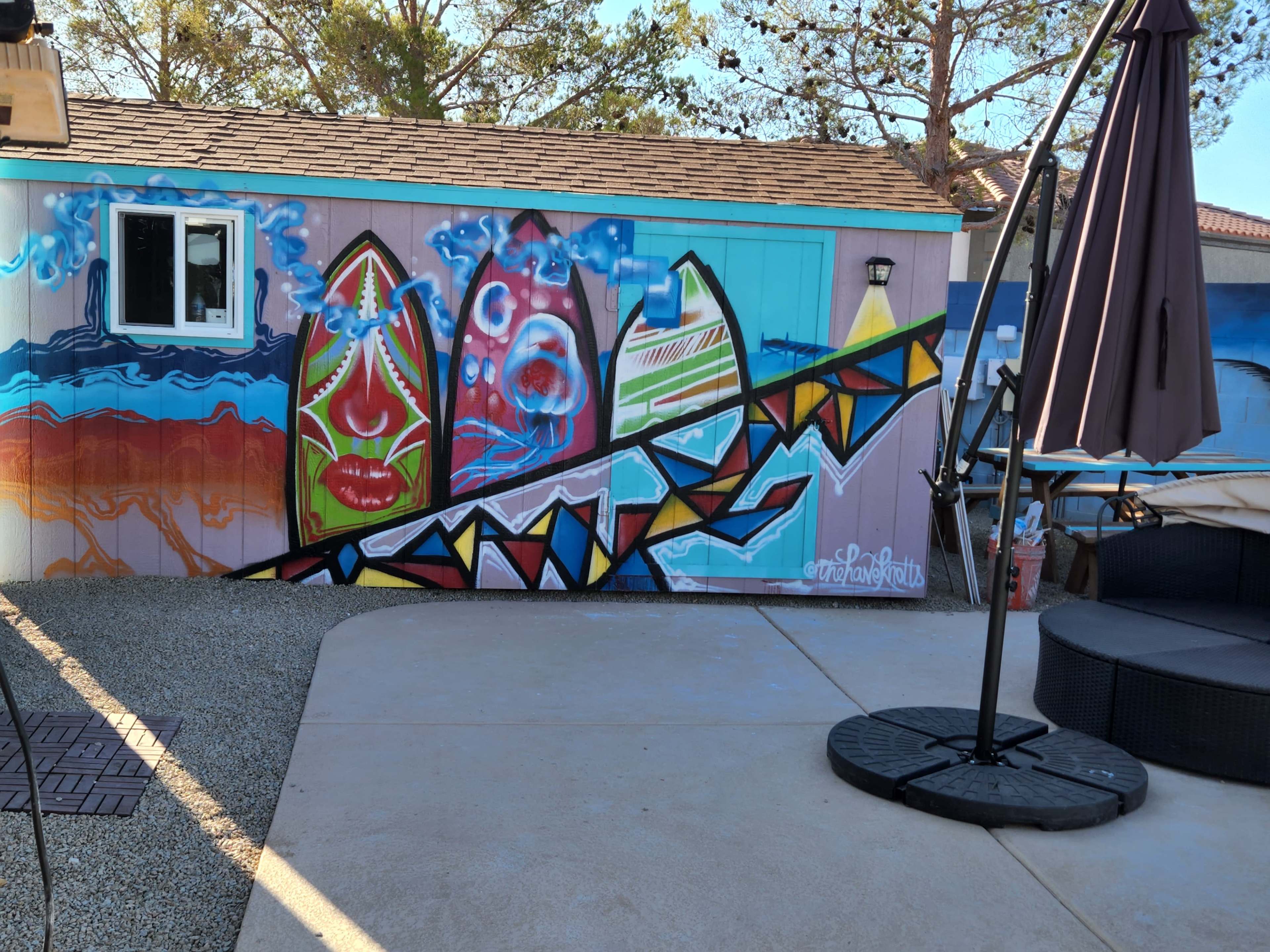 A colorful mural on a pink wall depicting abstract shapes and patterns, alongside a shaded area with a patio umbrella and a circular seating arrangement.