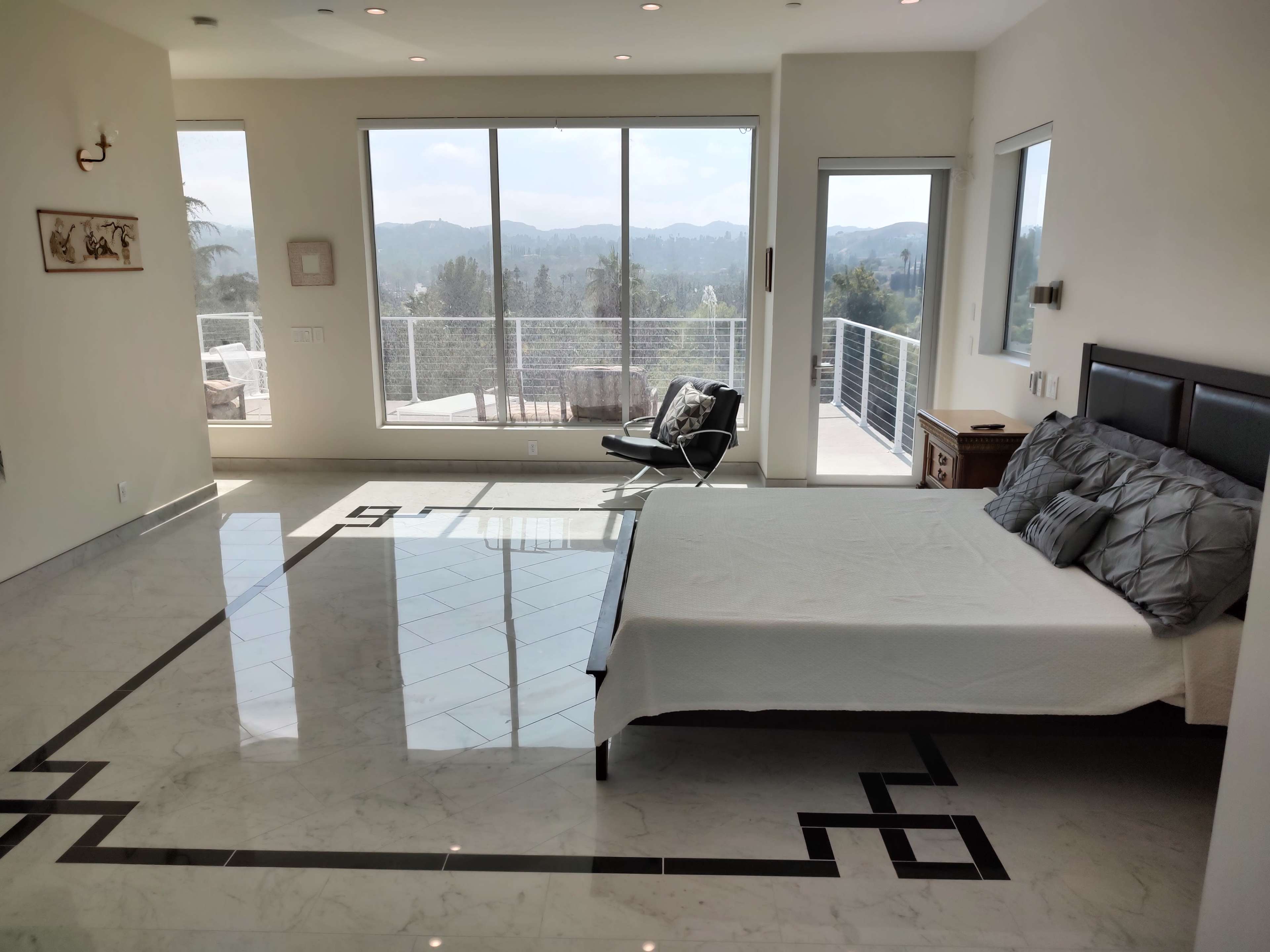 A modern bedroom with a large window that provides a view of mountains, featuring a bed, a chair, and marble flooring with a geometric design.