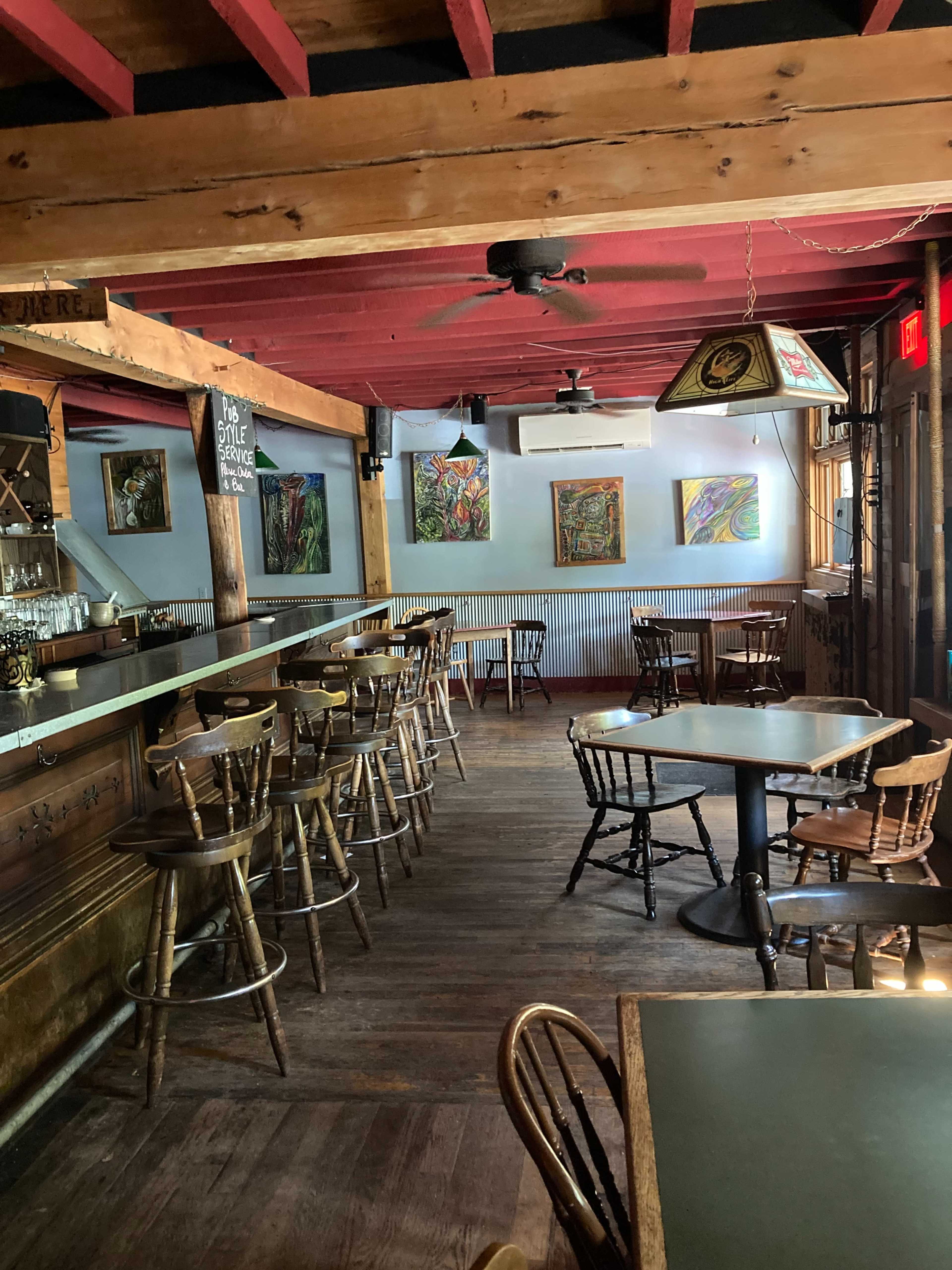 The interior of a restaurant with wooden floors, a bar area, and several tables and chairs arranged throughout, featuring colorful artwork on the walls.