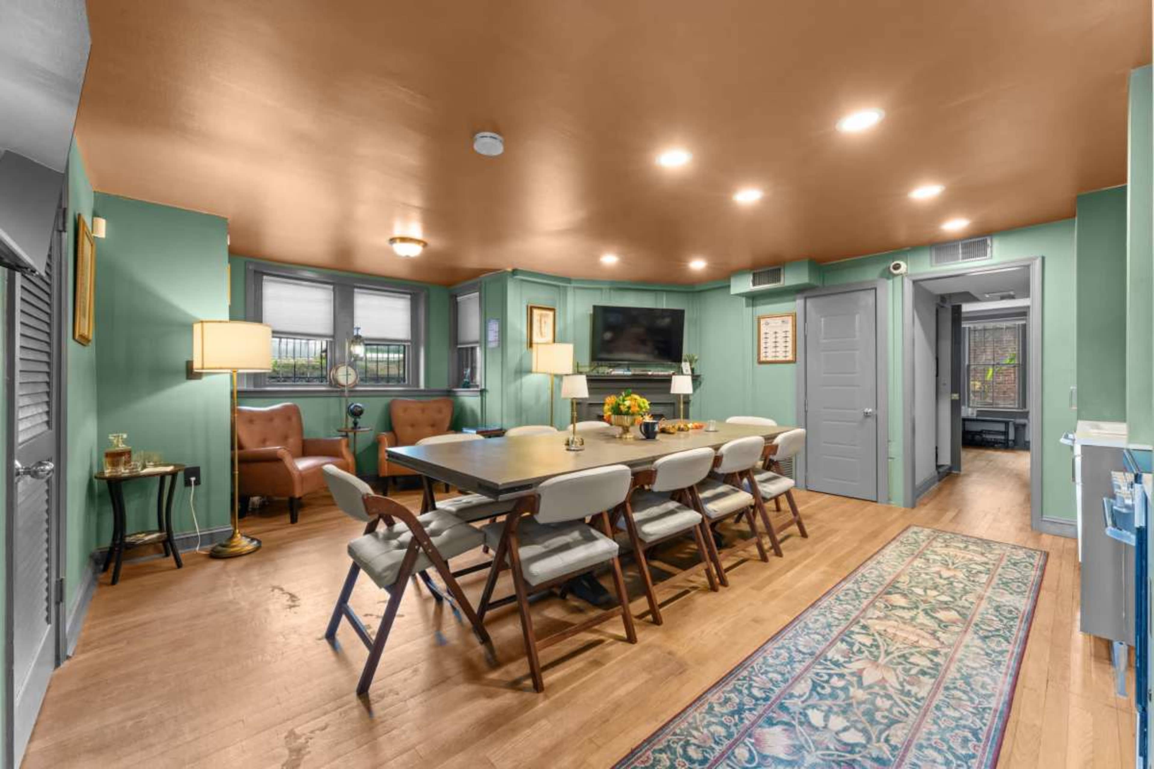 The image shows a spacious dining area with a large table surrounded by chairs, complemented by a seating area and a television mounted on the wall.