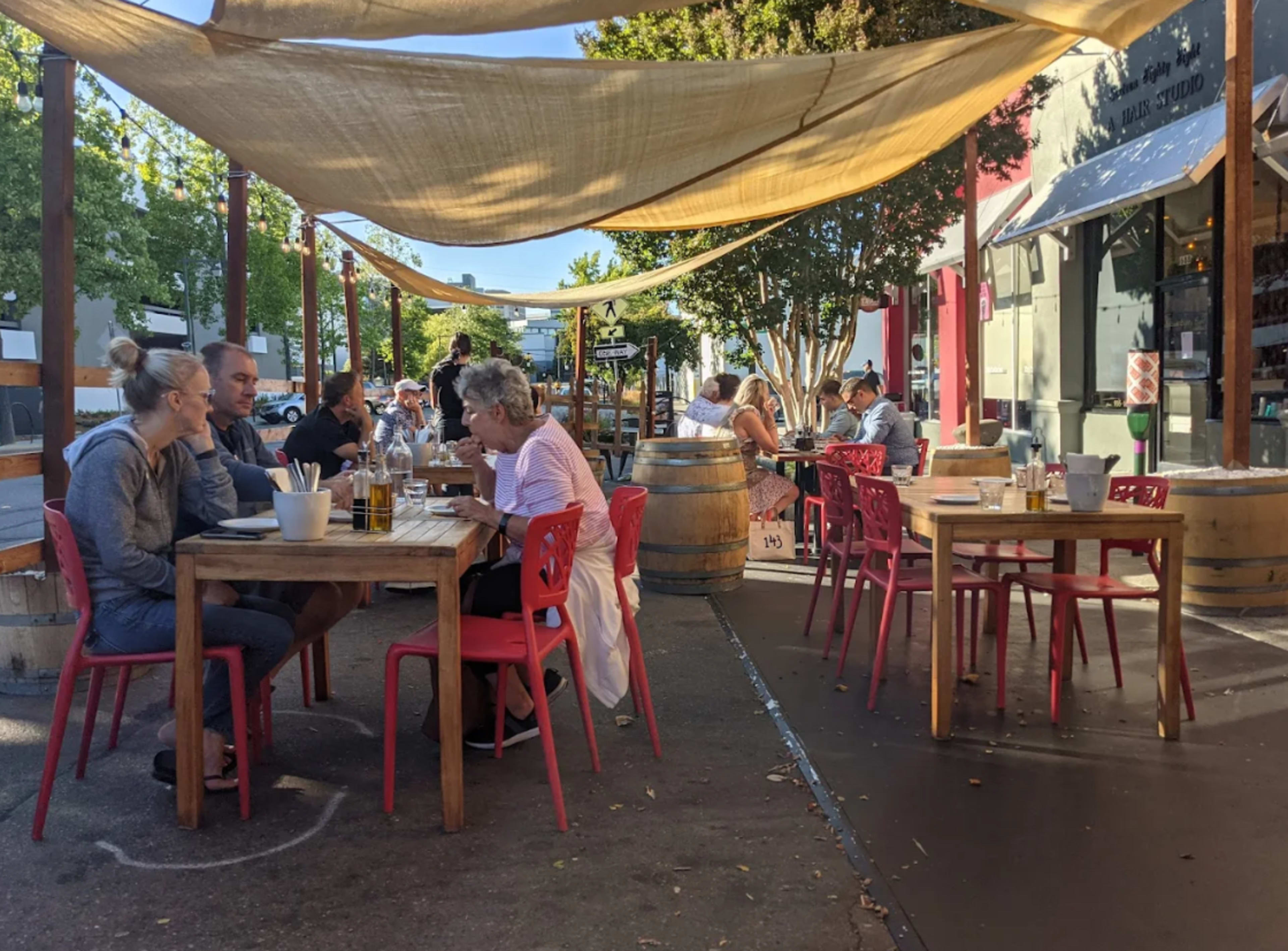 A busy outdoor dining area features people seated at wooden tables under a canopy, with some patrons eating and others engaging in conversation.