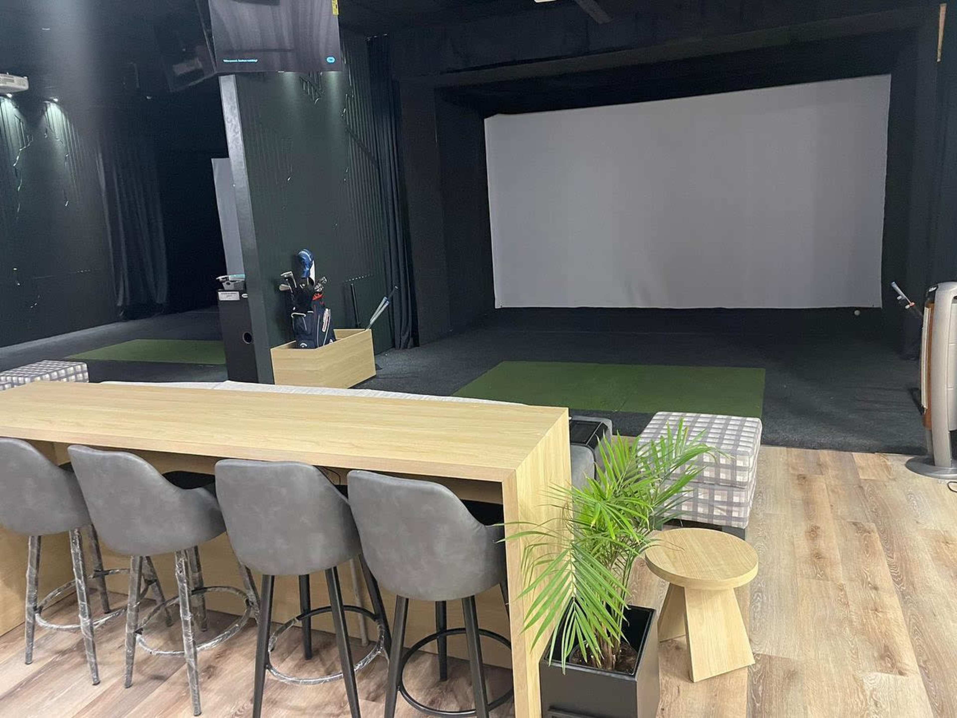 A modern indoor golfing facility featuring a bar area, seating, and a large projection screen at the front.