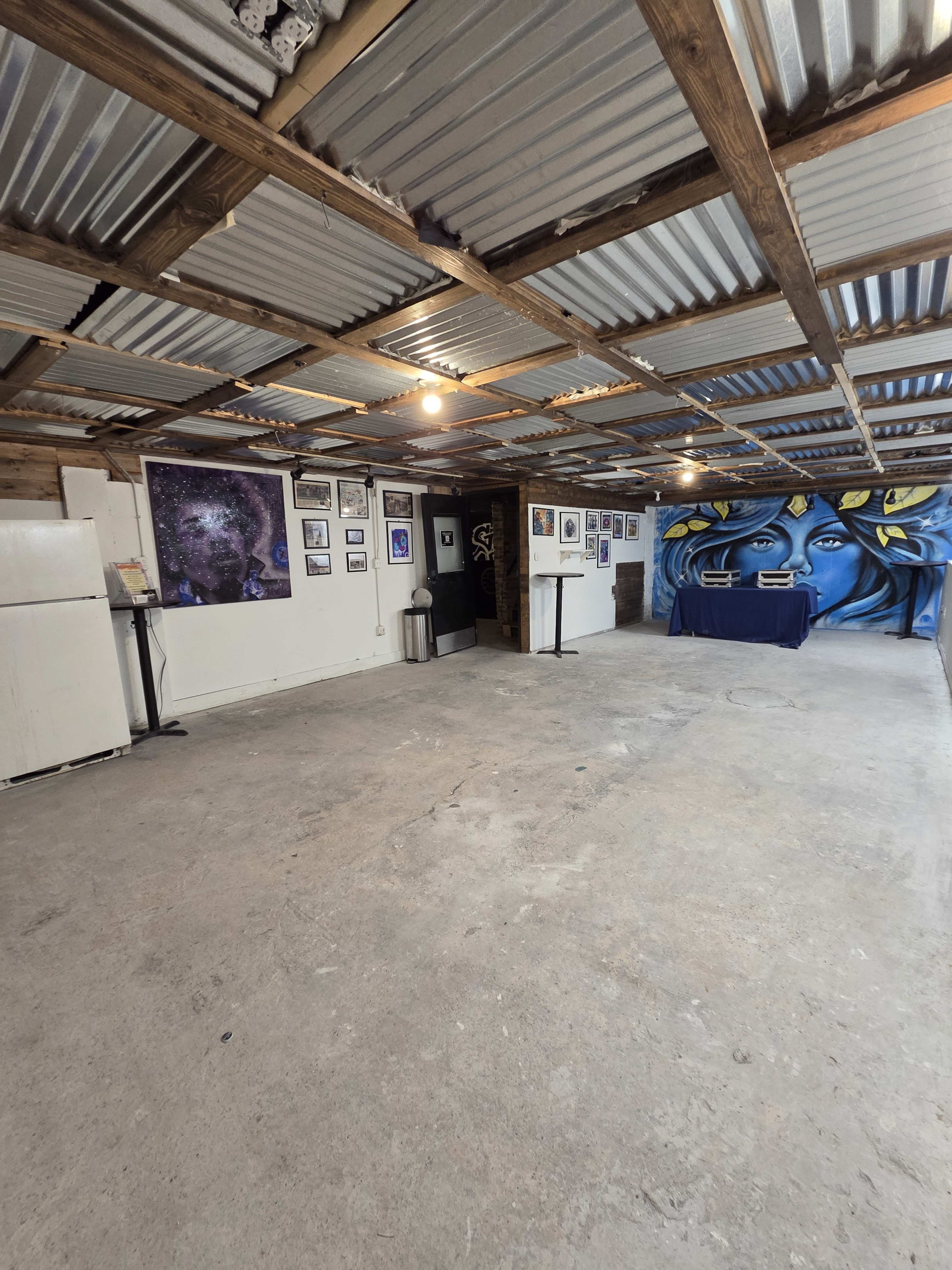 The interior of a space features exposed metal roofing, a series of framed artworks on the walls, a table covered with a cloth, and a large mural of a face on one wall.