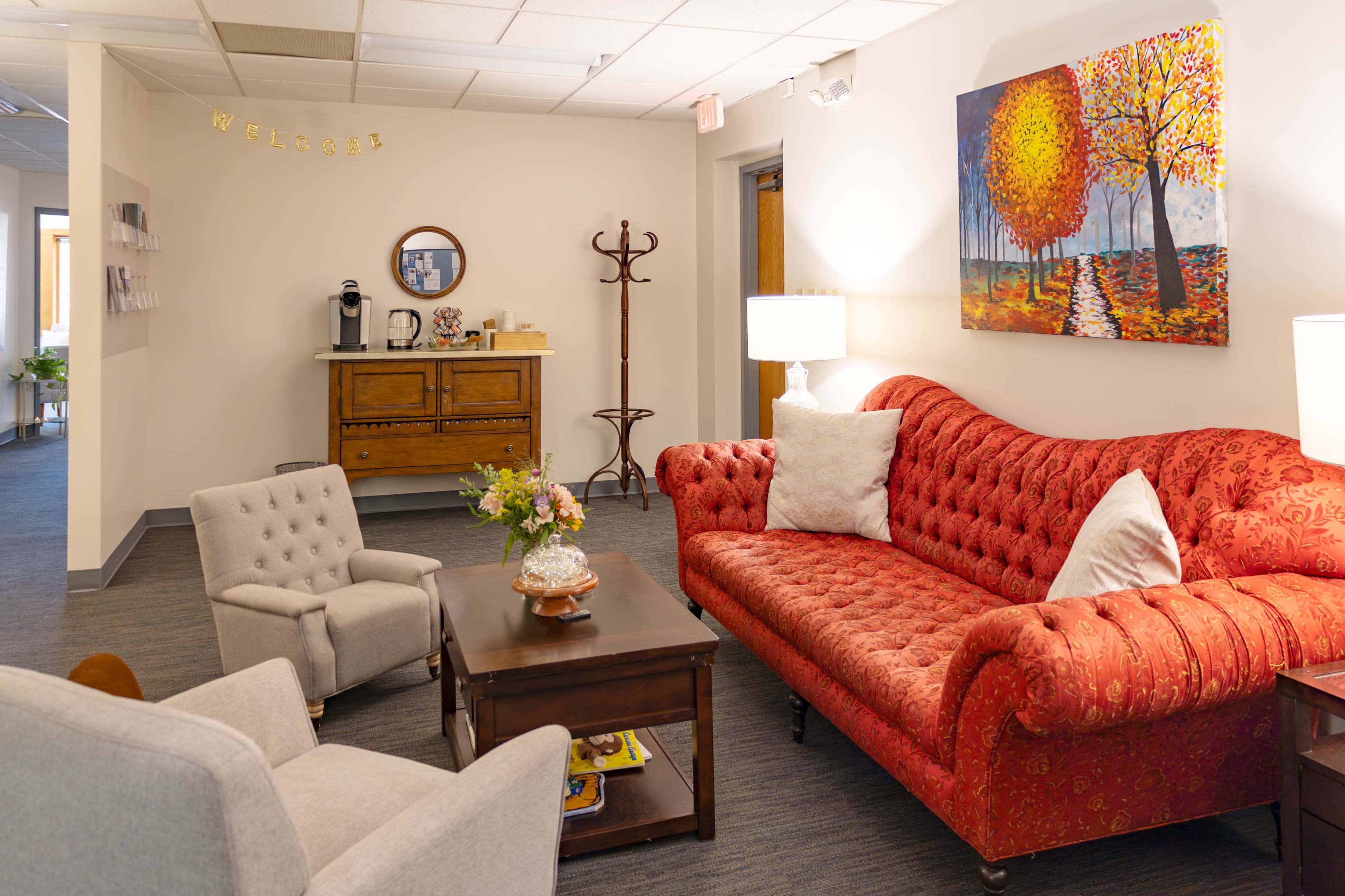 The image shows a cozy waiting area featuring a red tufted sofa, a pair of light gray armchairs, a wooden coffee table, and a sideboard with a coffee maker against the wall.