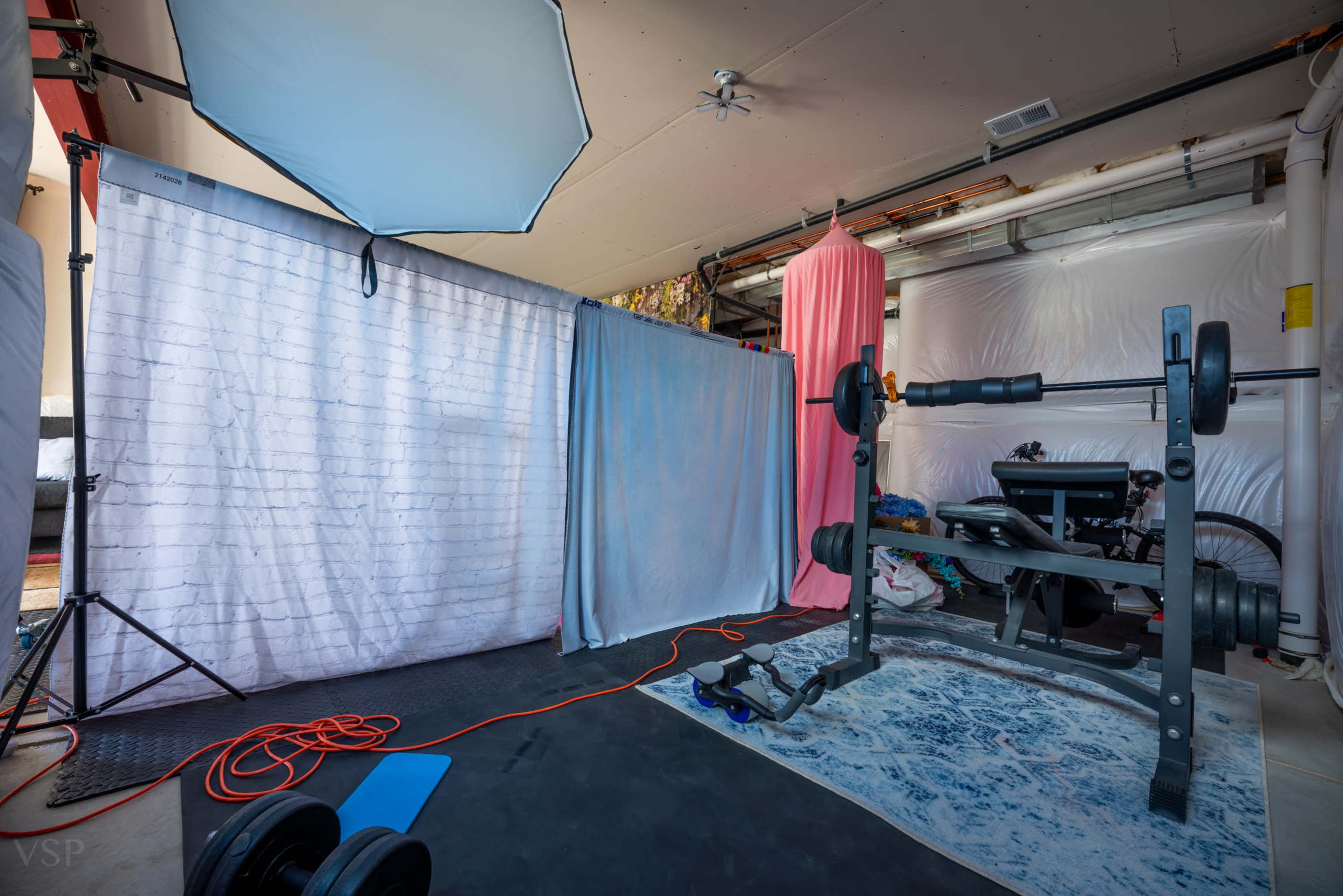 The image shows a makeshift gym setup in a garage, featuring a barbell on a weight rack, exercise equipment, and backdrops made of fabric.