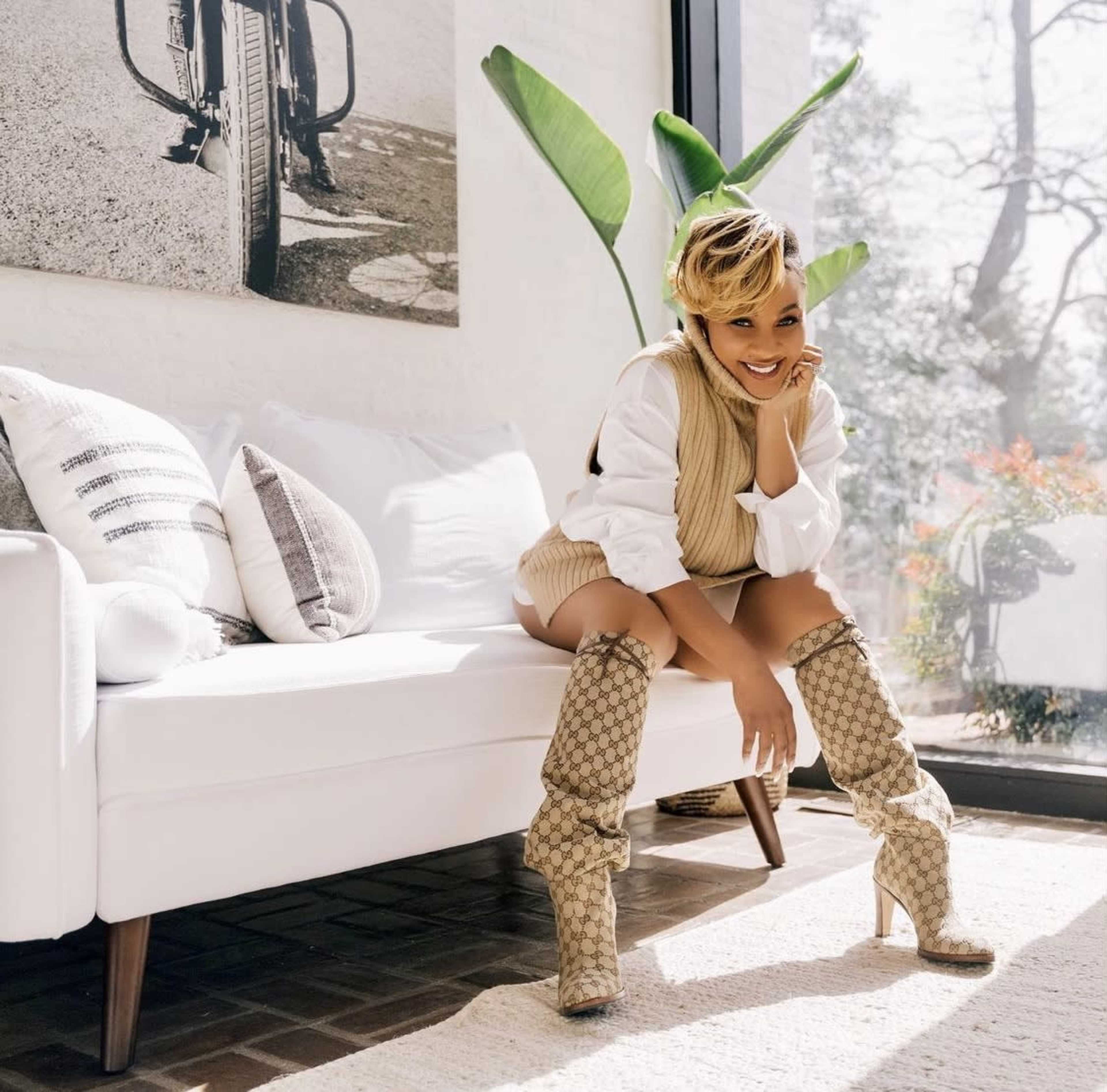 A woman in a beige sweater vest and knee-high patterned boots sits on a white sofa in a bright room with plants and large windows.