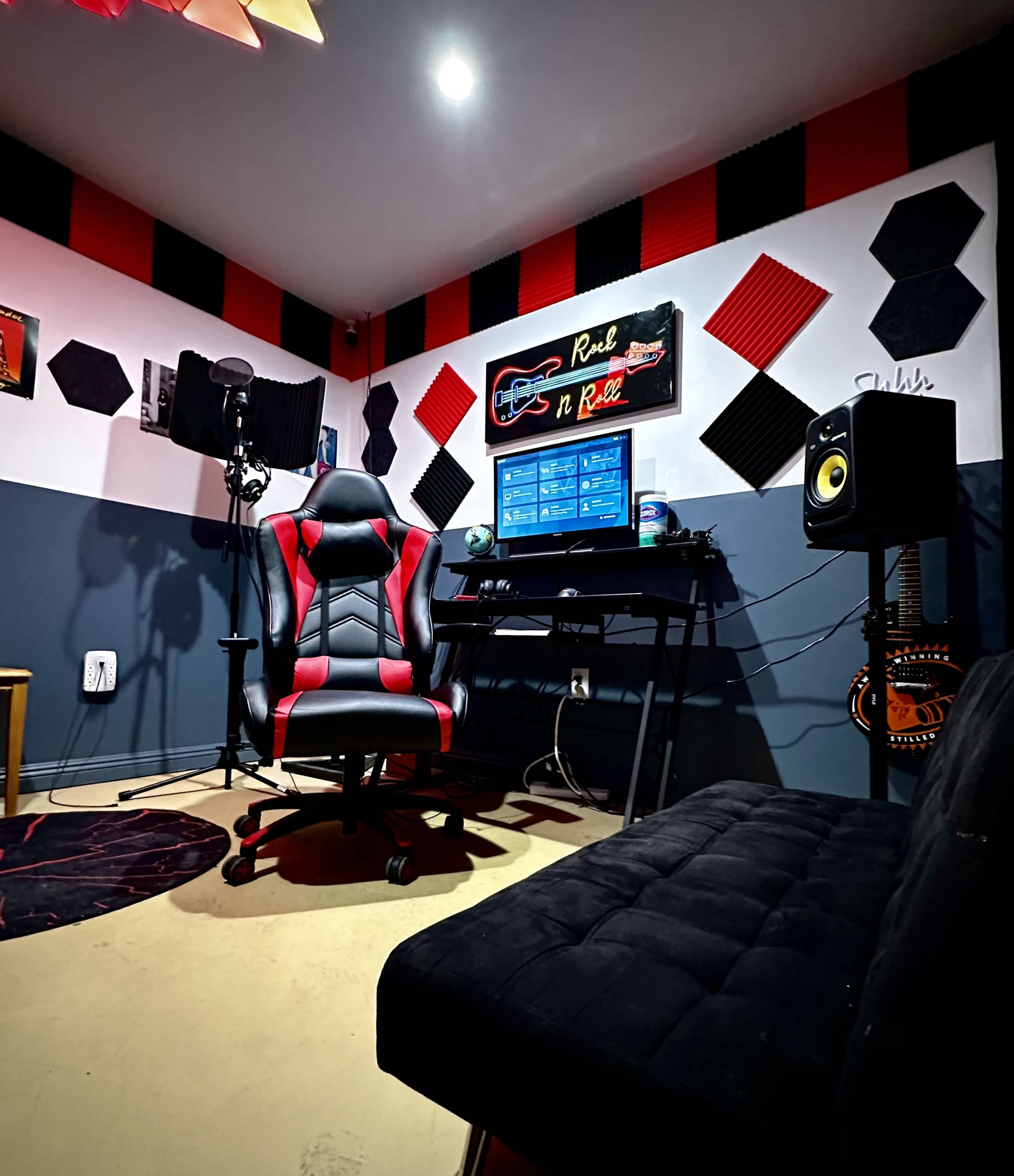 The image shows a modern recording studio with a desk, a computer monitor displaying audio software, a gaming chair, and soundproofing panels on the walls.