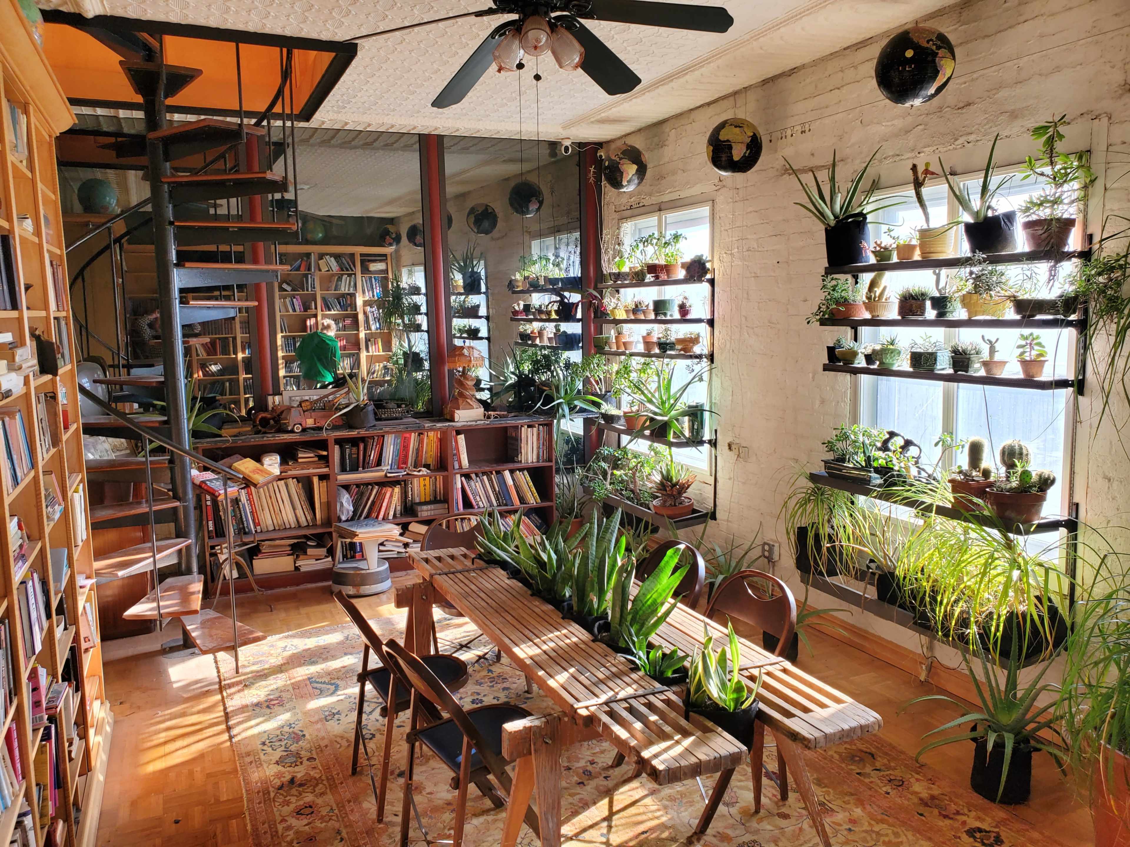 A cozy, plant-filled reading room with wooden shelves of books, a spiral staircase, and a wooden table surrounded by greenery.